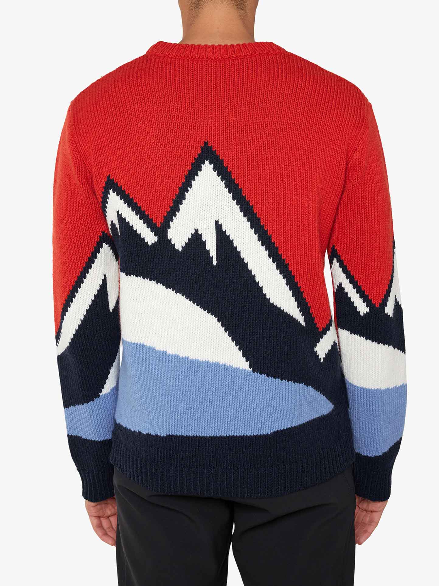 A person is wearing the Snowmountain Sweater Men by We Norwegians, a cozy cashmere blend featuring a bold mountain design on the back. The pattern showcases snow-capped peaks in black, white, and blue against a red background. The person stands with their back to the camera, highlighting the sweater's striking and comfortable look.