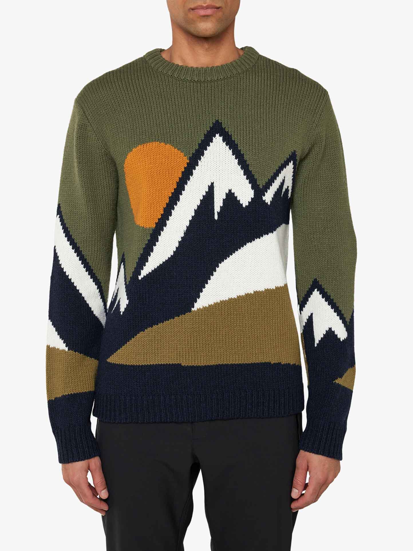 A person is wearing the We Norwegians Snowmountain Sweater Men, a green knitted cozy sweater featuring a mountain design with white peaks, a setting orange sun, and dark blue accents. Made from a soft cashmere mix, this sweater has a round neck and is paired with black pants. The image focuses on the upper body and showcases the detailed pattern of the sweater.