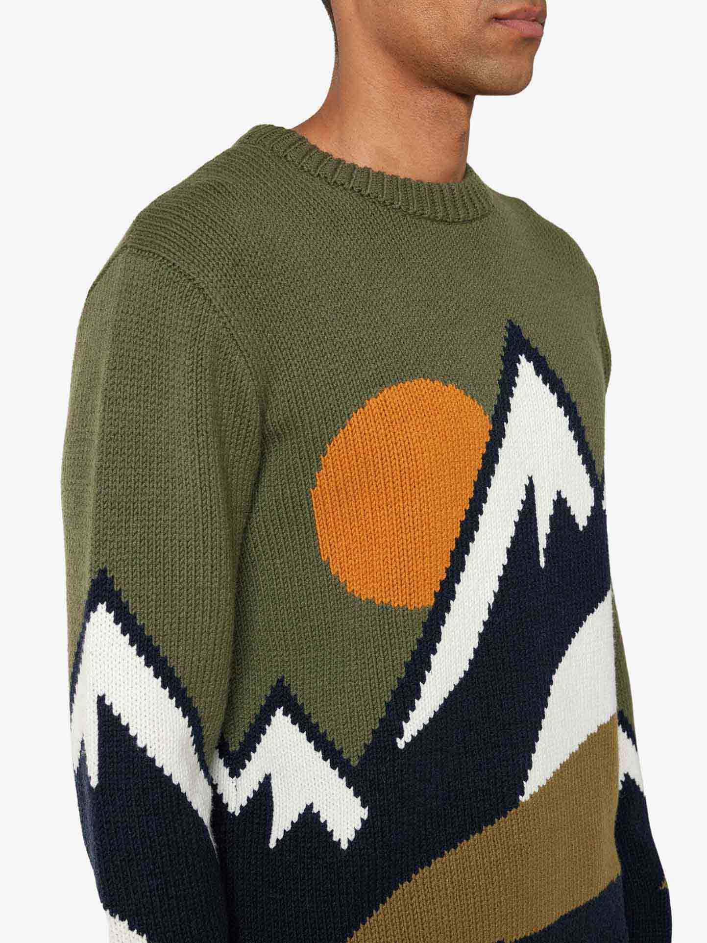 A person wearing the We Norwegians Snowmountain Sweater Men, a cozy green knitted sweater made from a soft cashmere mix, featuring a design of snow-capped mountains and an orange sun. The sweater has a ribbed neckline, and the person is shown from the shoulders to the lower face, with their left side visible. The background is plain white.