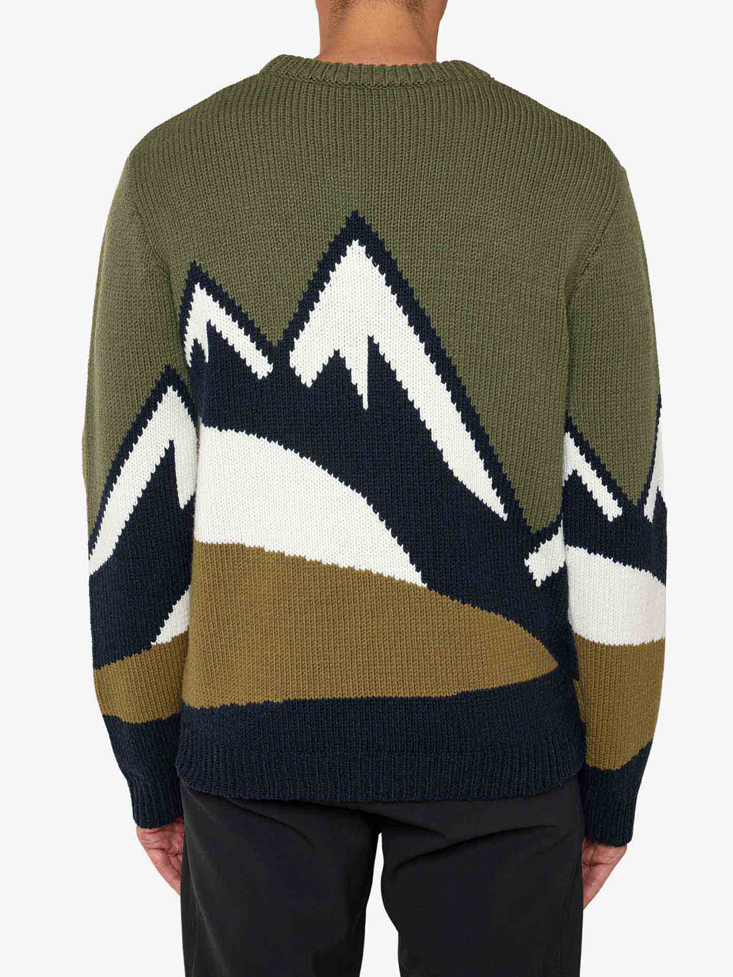 A person is wearing the We Norwegians Snowmountain Sweater Men, a cozy, green merino wool sweater featuring a mountain design on the back. The mountains are depicted in shades of white, navy blue, and brown. The person stands with their back to the camera.