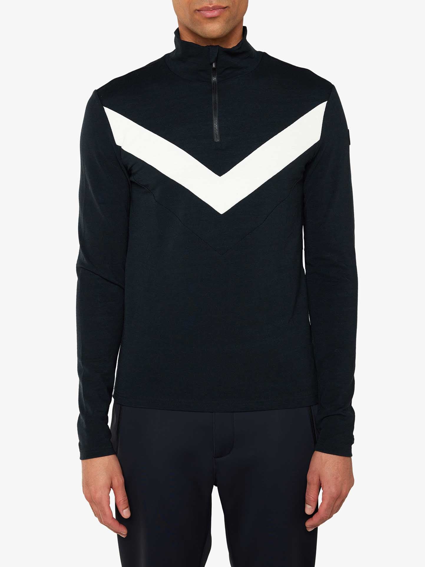 A person wearing a We Norwegians Voss Chevron Zipup Men in black, featuring a white chevron design on the chest and a half-zip collar, stands against a plain white background, exuding retro vibes. The pullover, crafted from superfine Merino wool, is paired with black pants.