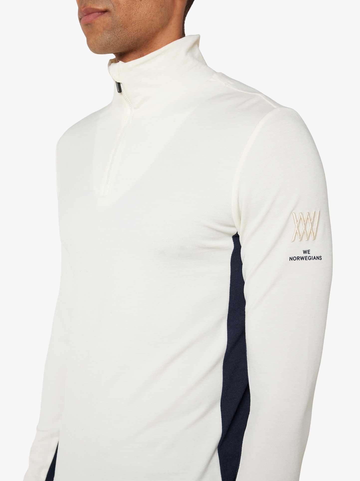 A person wearing a We Norwegians Voss 1/4-Zip Men in white with black color block panels. This top, crafted from superfine merino wool, features the brand name "We Norwegians" and a logo on the left sleeve. The zipper is partially open, creating a casual and sporty look.