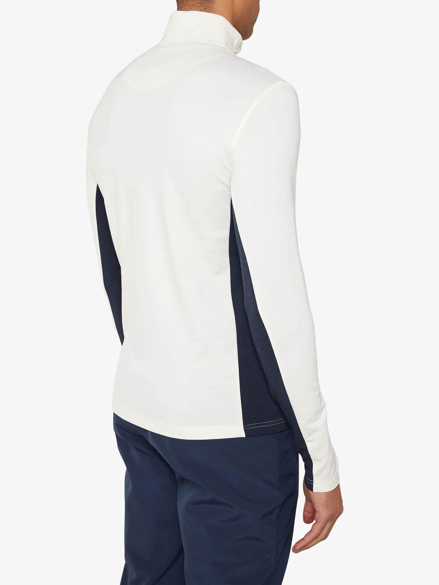 A person is seen from the back wearing the Voss 1/4-Zip Men shirt by We Norwegians, a white long-sleeve shirt with dark color block panels and dark navy-blue pants. The high-collared shirt, potentially crafted from superfine Merino wool, is worn casually with the person's right arm relaxed at their side against a plain white background.