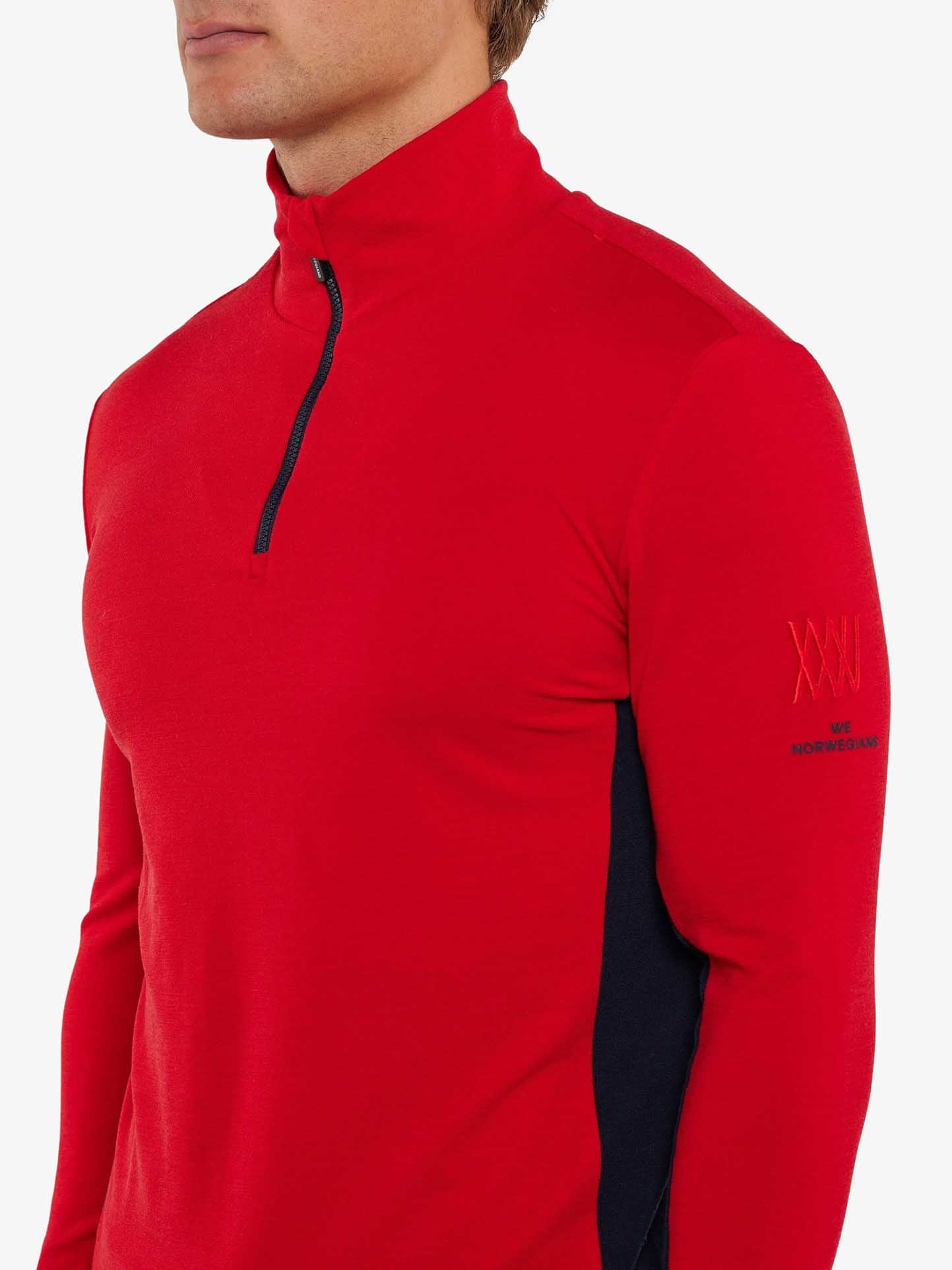 A person is wearing a red long-sleeve Voss 1/4-Zip pullover from We Norwegians, crafted from Superfine Merino wool. It features a black zipper on the chest and color block panels on the sides. The pullover has subtle branding on the left arm, and the person's face is out of the frame.