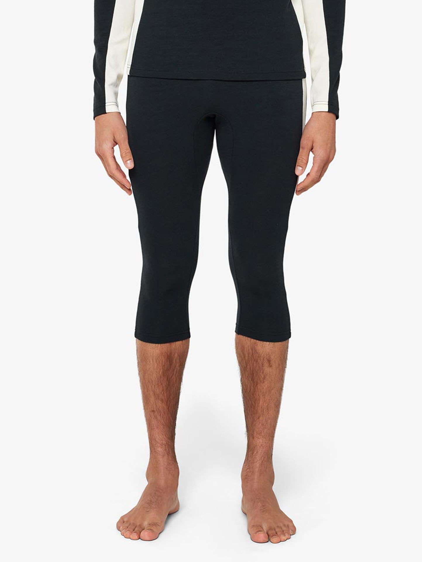 A person stands barefoot on a plain background, wearing the "Voss Long Johns Bottoms Men" by We Norwegians, which are tight-fit black cropped athletic leggings ending below the knees. They are also wearing a matching long-sleeve top featuring white side panels on the sleeves.