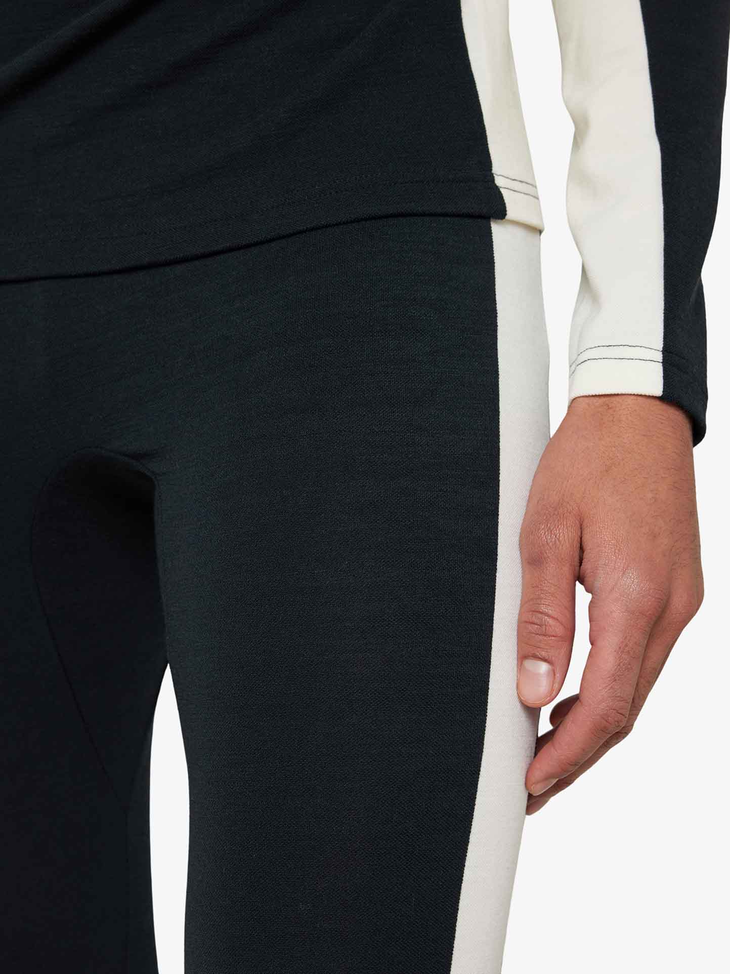 A close-up of a person wearing We Norwegians' Voss Long Johns Bottoms Men, featuring a two-toned Merino wool design. The left half of the bottoms is black, while the right half is white. Only the lower torso and right arm are visible, with the person's right hand resting by their side.