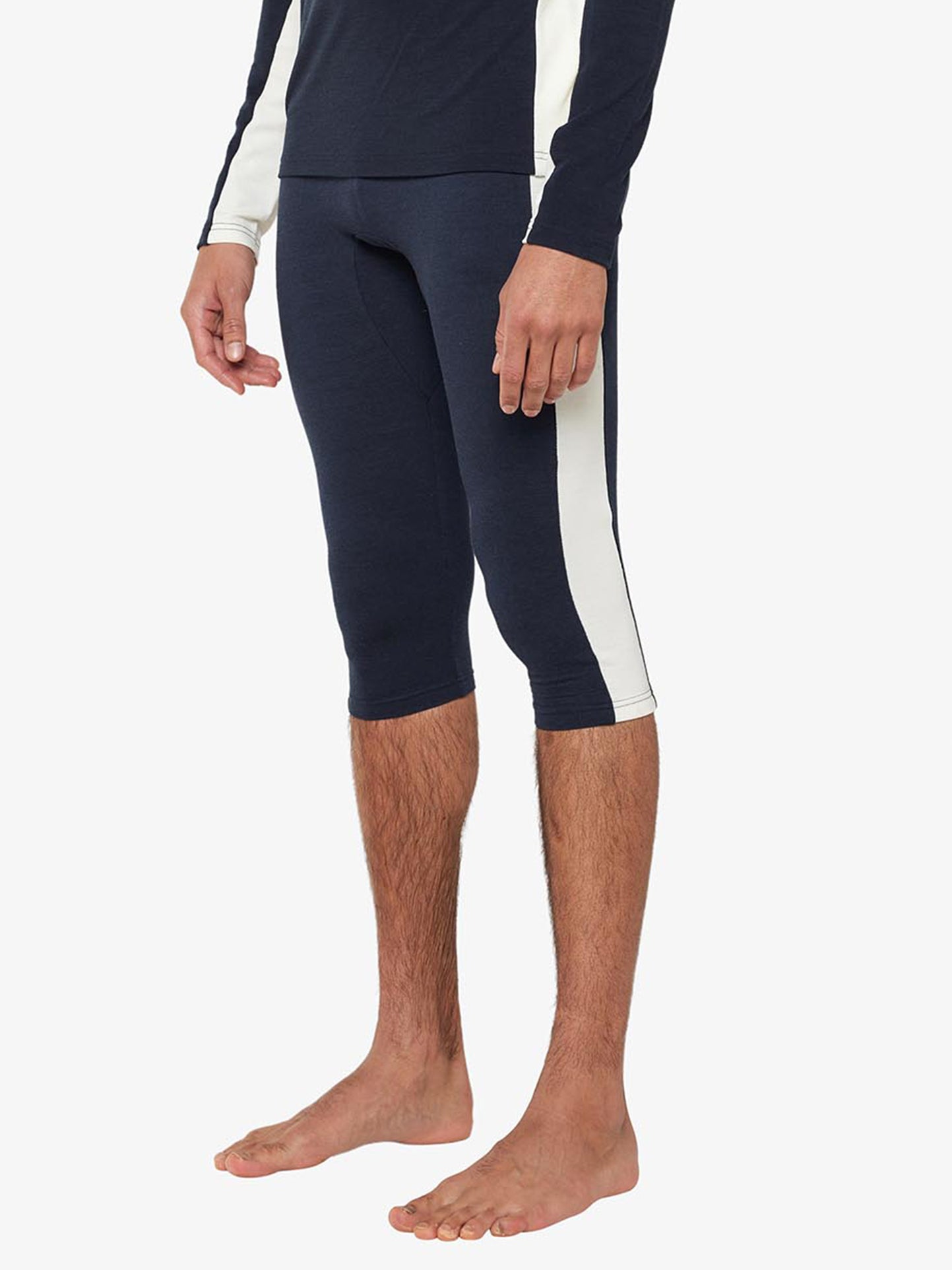 An individual is wearing the Voss Long Johns Bottoms Men from We Norwegians, which are dark-colored, three-quarter length long johns featuring a wide vertical white stripe on the outer side. They are also donning a matching top with white stripes down the sleeves. Only the lower half of their body is visible.