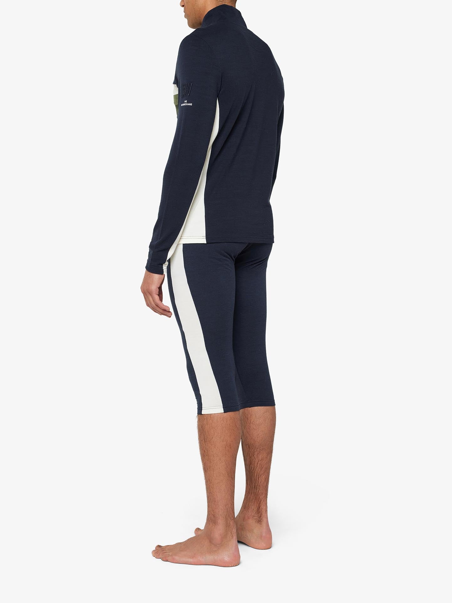 A person is standing barefoot, dressed in the navy blue We Norwegians Ski 1/4-Zip Men outfit with cream-colored side panels fabricated from superfine Merino wool. The long-sleeved top and three-quarter length leggings create a retro-inspired base layer look as the person stands with a slight side profile, facing away from the camera.