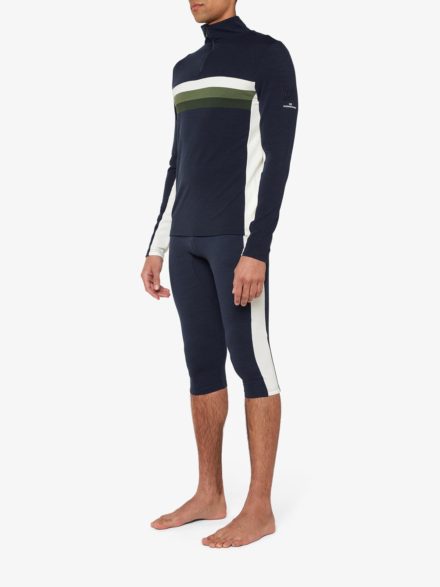 A person is standing barefoot, dressed in the retro-inspired We Norwegians Ski 1/4-Zip Men base layer made from superfine Merino wool. The dark blue athletic outfit features a long-sleeve top with white and olive green horizontal stripes, complemented by three-quarter length leggings adorned with white vertical stripes down the sides.