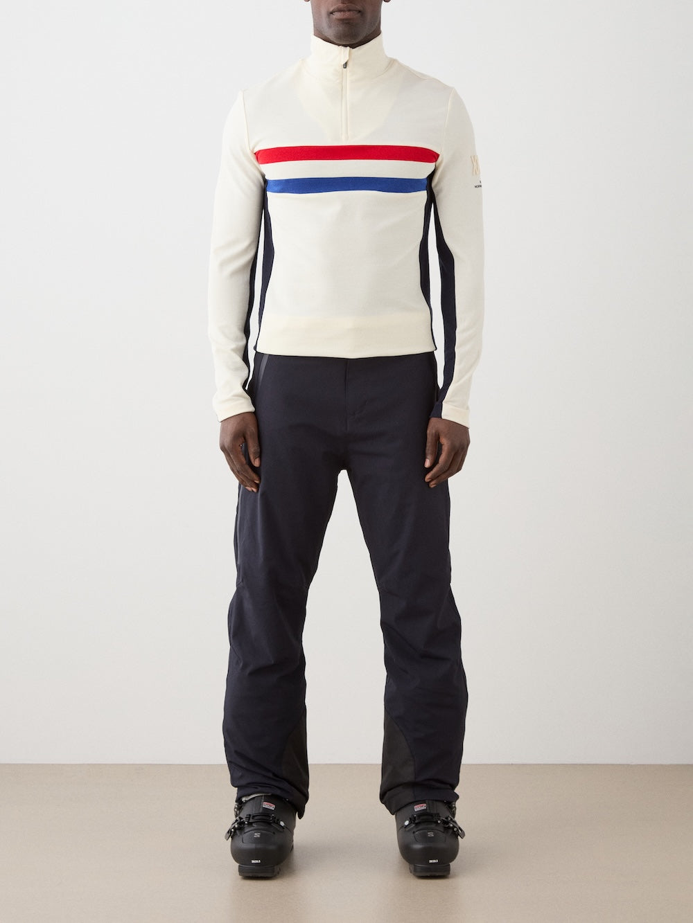 A person stands facing forward wearing the We Norwegians Ski 1/4-Zip Men sweater with red and blue chest stripes, paired with black pants and black ski boots against a plain background.