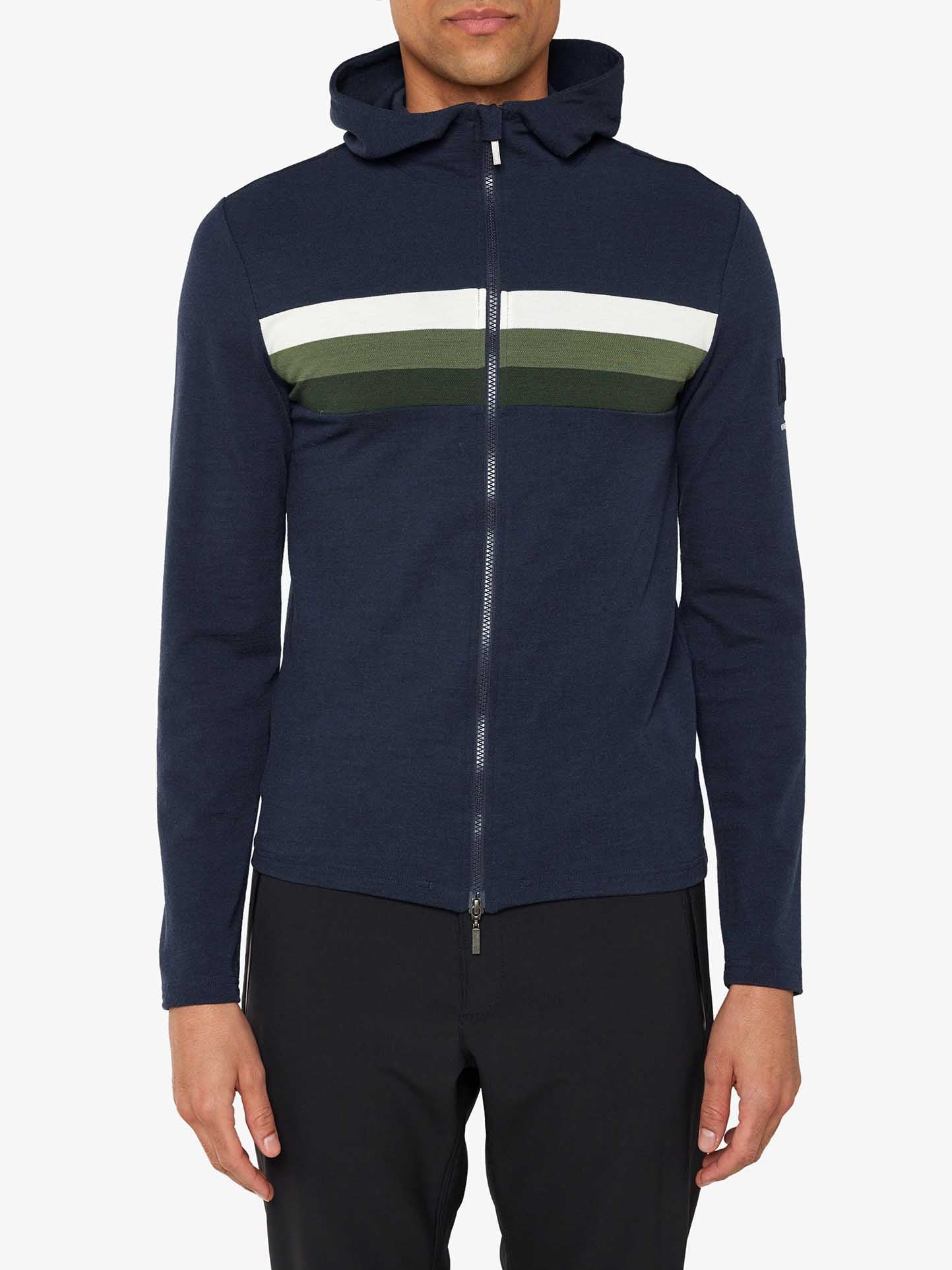 A person wearing the We Norwegians Ski Fullzip Hoodie Men, featuring a navy blue, retro color block design with a hood and two horizontal stripes—one white and one green—made from merino wool. The hoodie is paired with black pants against a plain white background.