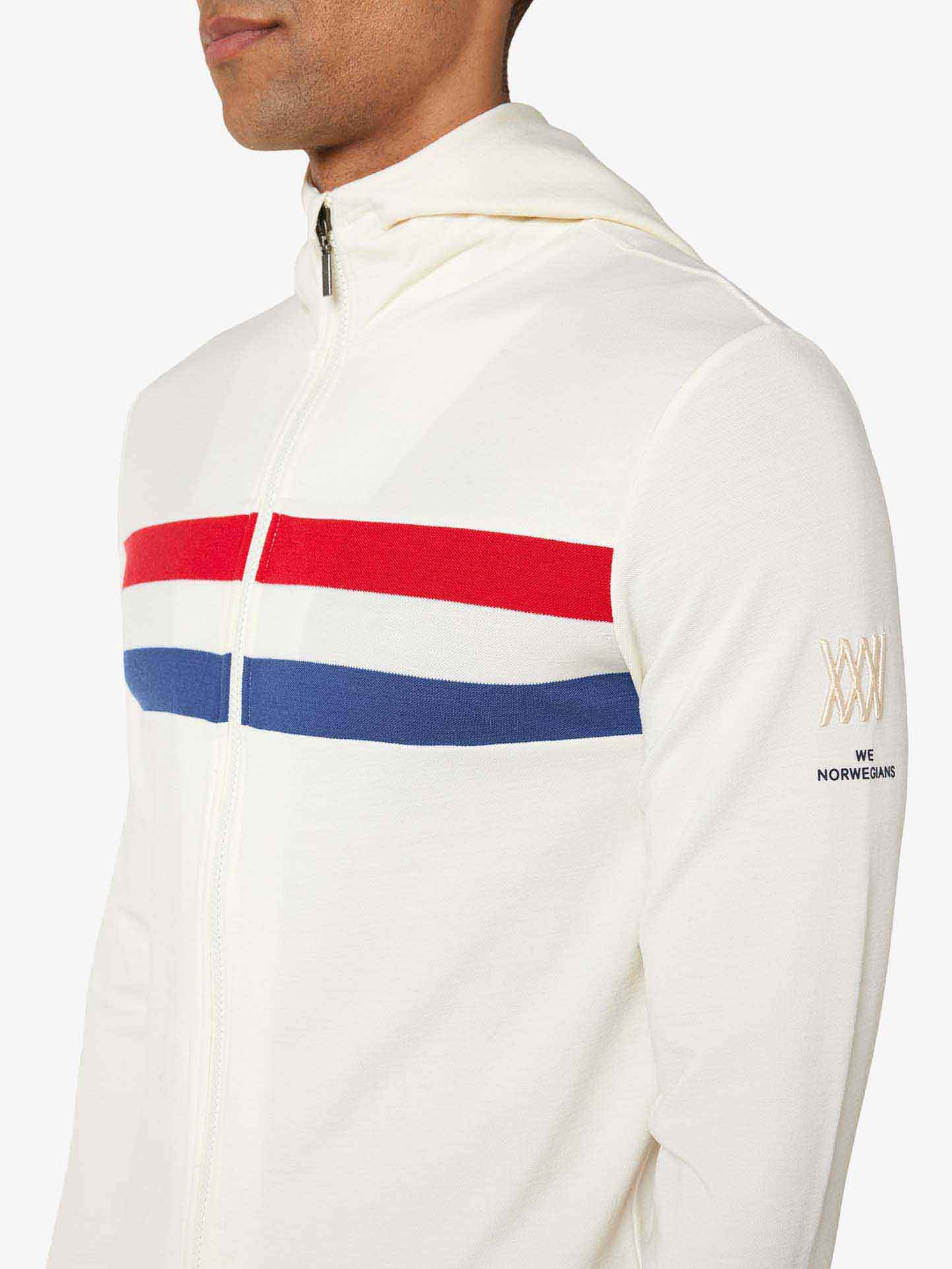 A person wearing the "Ski Fullzip Hoodie Men" from We Norwegians, which features horizontal red and blue stripes across the chest on a white background. The left sleeve has "We Norwegians" embroidered near the wrist. Made from merino wool, this retro color block design captures the upper torso and part of the face.