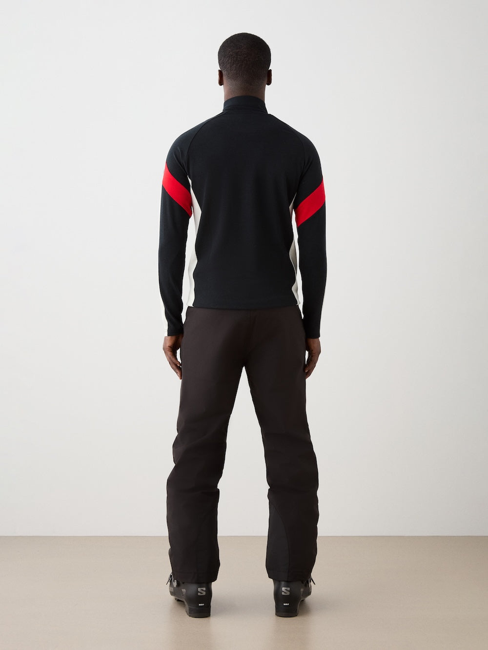 The We Norwegians Zermatt Zipup Men is a Merino wool ski sweater with a half-zip collar, black body, cream sleeves with black inner arms, diagonal red stripes, and a circular patch on the left sleeve | Back