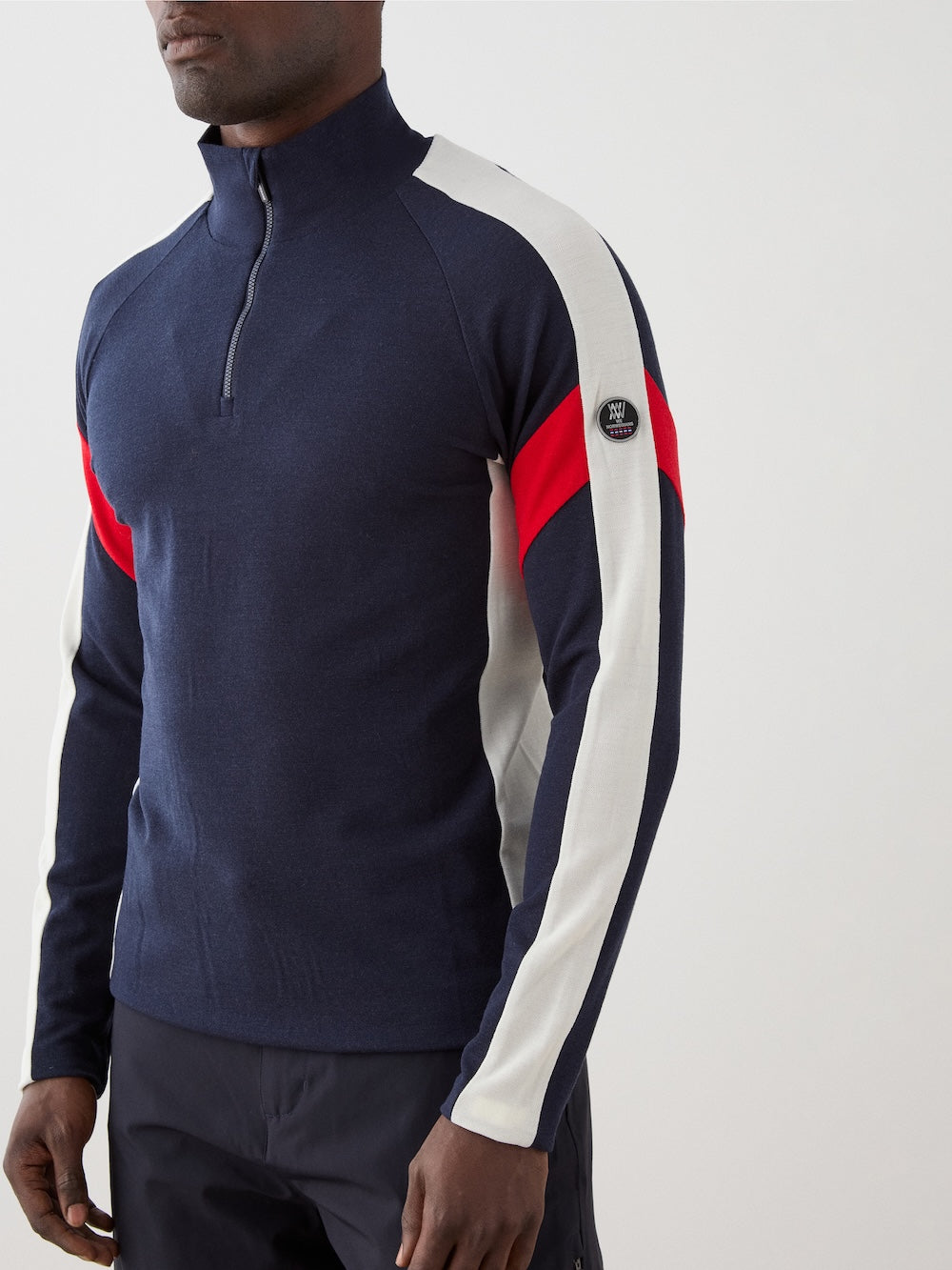 The We Norwegians Zermatt Zipup Men is a navy long-sleeve ski sweater with a quarter-zip, white shoulder and sleeve panels, red diagonal stripes on the upper sleeves, and a small circular logo patch on the left upper arm | Close-up