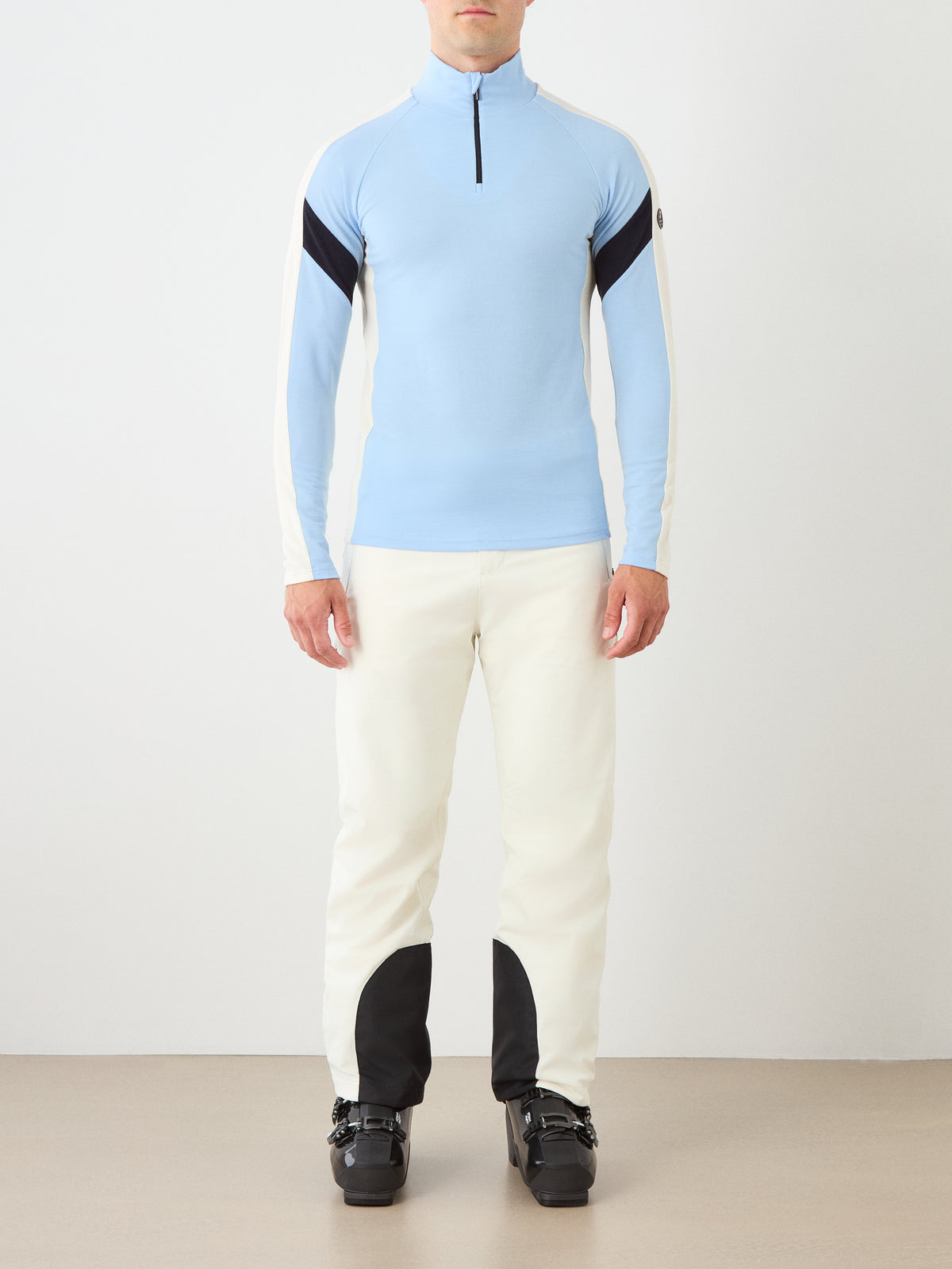 A person stands facing forward against a plain light background wearing the We Norwegians Zermatt Zipup Men in light blue and white, crafted from Merino wool, paired with white pants featuring black knee patches and black ski boots.