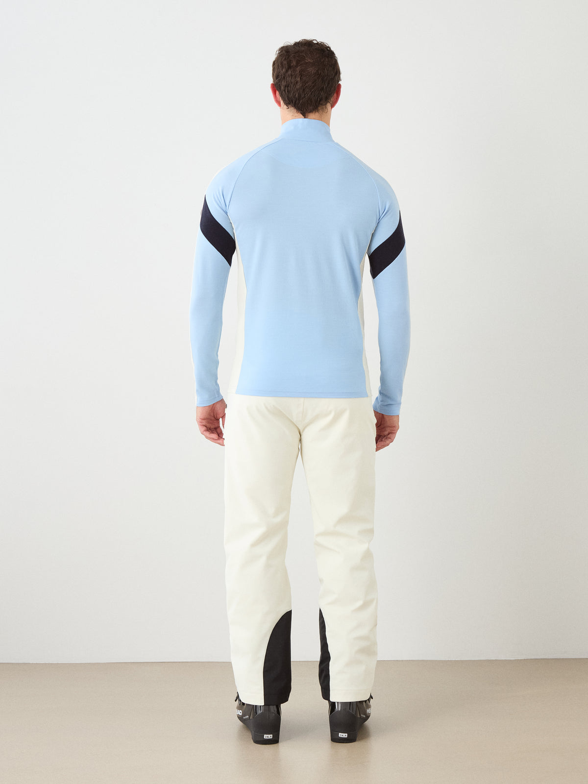 A man stands facing away from the camera, wearing the We Norwegians Zermatt Zipup Men in light blue—a soft Merino wool long-sleeve top from the menswear collection—paired with white pants featuring black calf patches and black shoes, set against a plain indoor background.