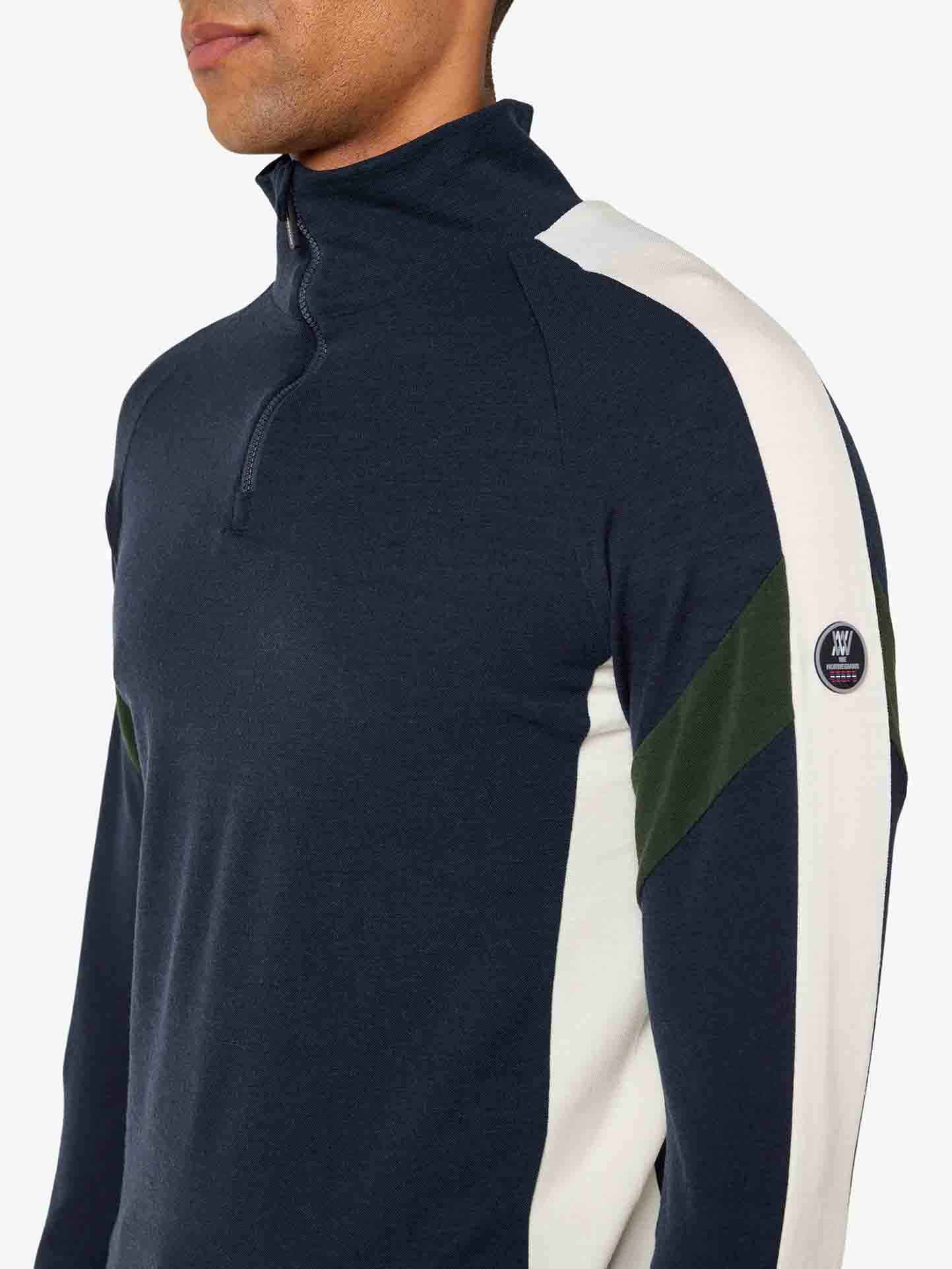 A person is wearing a dark, long-sleeved Zermatt Zipup Men from We Norwegians, featuring white and green stripes on the sleeves and a small circular logo on the left sleeve. Made from superfine Merino wool, it’s part of a sleek ski ensemble against a plain white background.