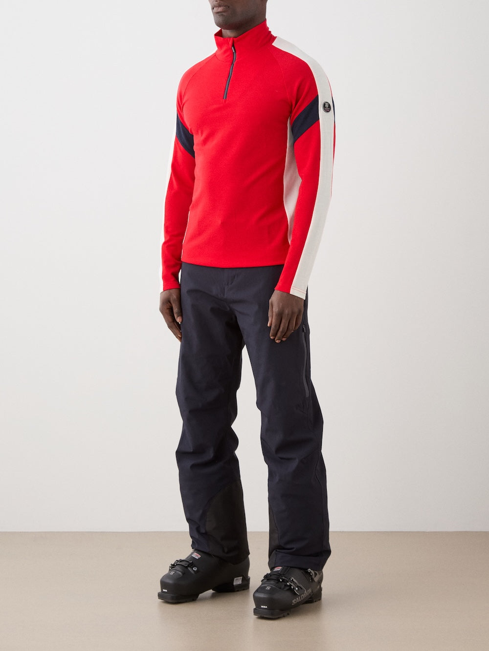 The We Norwegians Zermatt Zipup Men is a red long-sleeve ski sweater with a half-zip front, white sleeve panels, black diagonal arm stripes, and a circular logo patch on the upper left sleeve | Front