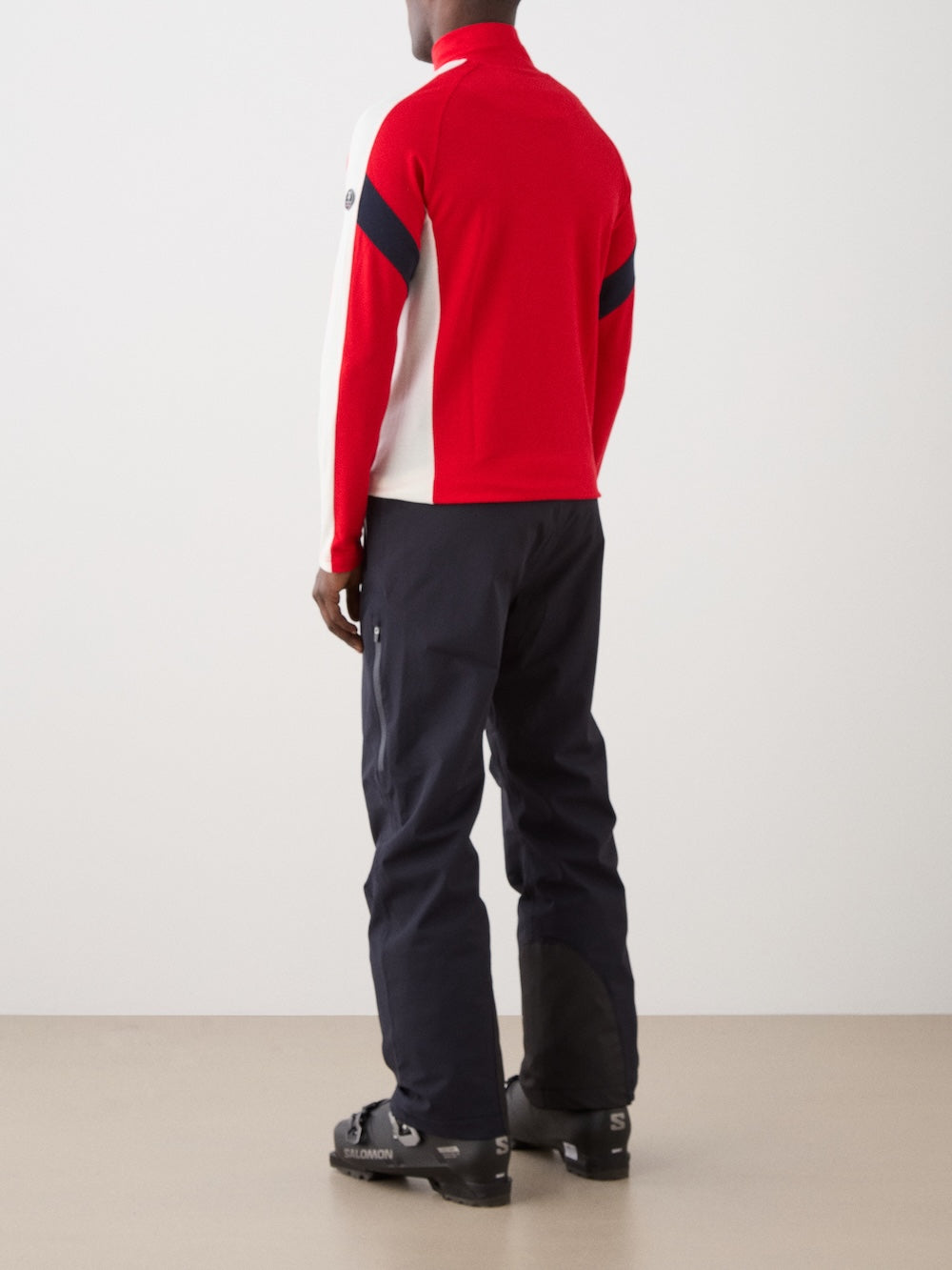 The We Norwegians Zermatt Zipup Men is a red long-sleeve ski sweater with a half-zip front, white sleeve panels, black diagonal arm stripes, and a circular logo patch on the upper left sleeve | Back