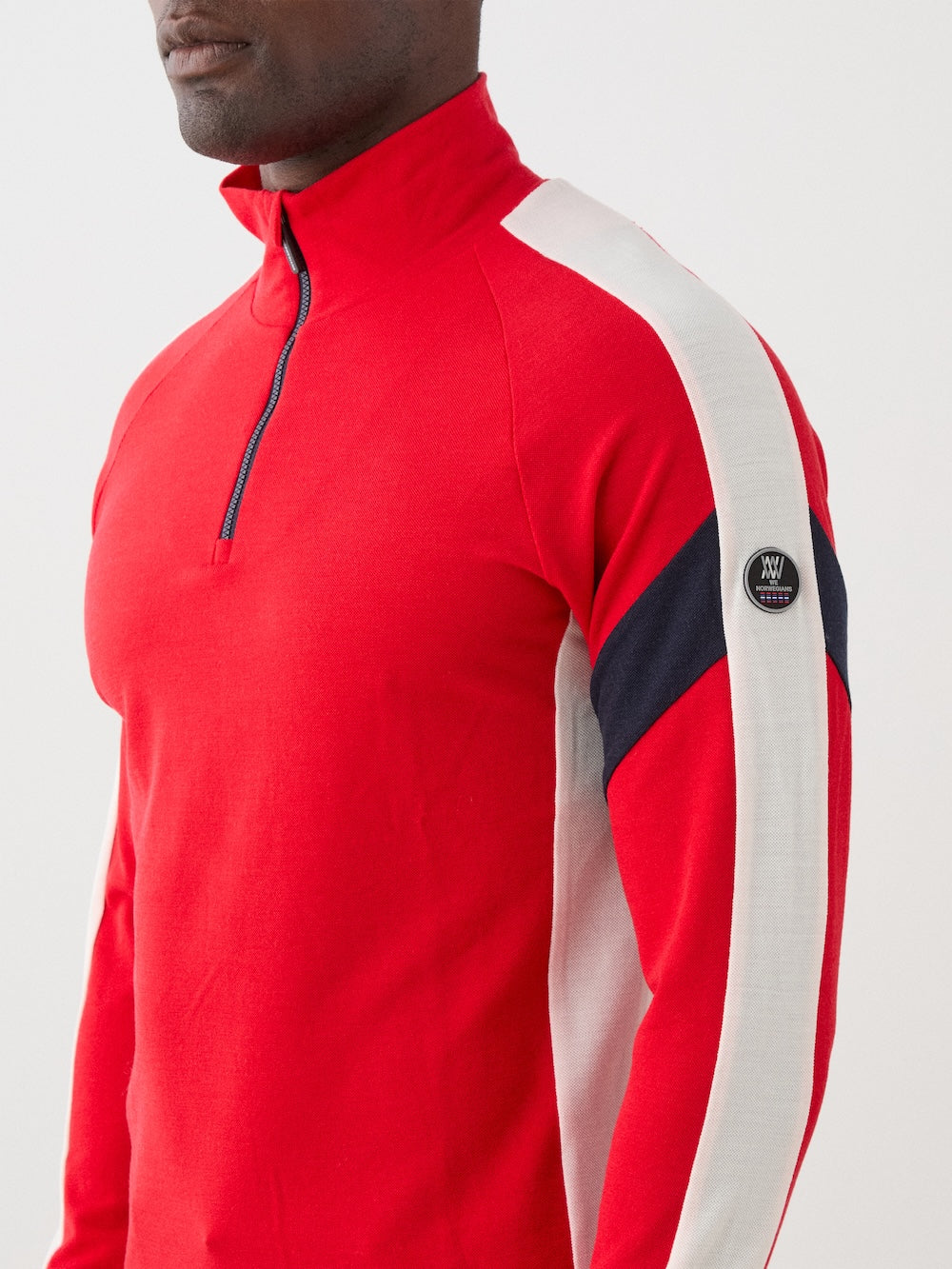 The We Norwegians Zermatt Zipup Men is a red long-sleeve ski sweater with a half-zip front, white sleeve panels, black diagonal arm stripes, and a circular logo patch on the upper left sleeve | Close-up