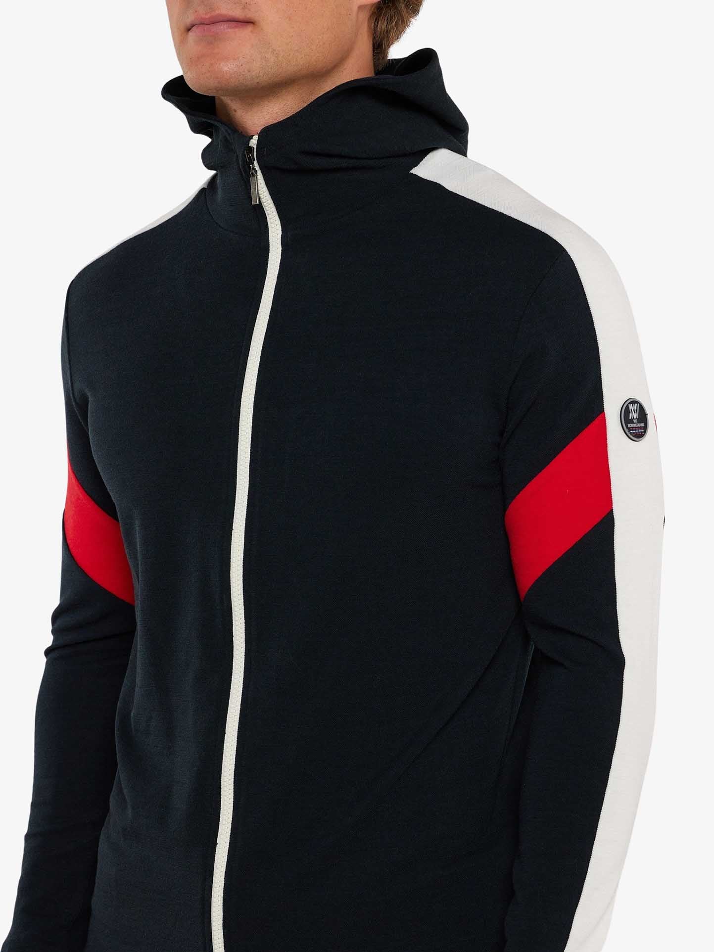 A person is wearing a black and white Zermatt Fullzip Hoodie Men from We Norwegians, featuring a high collar. Made from superfine Merino wool, the jacket has red diagonal stripes on the sleeves and a small round logo on the upper sleeve. The image shows the upper half of the person's body with part of their face slightly cut off above the nose.