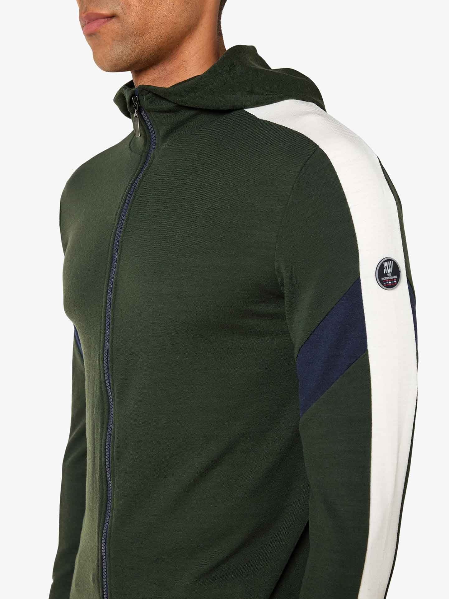 A person wearing a dark green Zermatt Fullzip Hoodie Men by We Norwegians, with white and dark blue accents on the sleeves. The athletic fit hoodie, crafted from Superfine Merino wool, features a hood and a small circular logo patch on the upper arm. The person is shown from the side, facing slightly away from the camera.