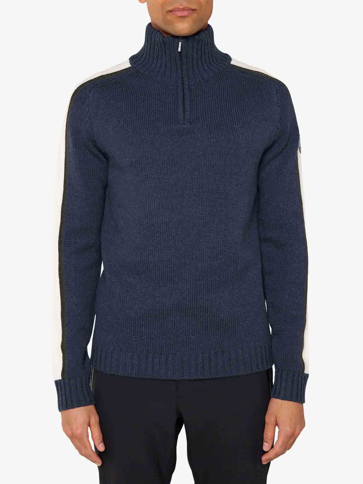 A person wearing a dark blue, high-neck Zermatt Sweater Men by We Norwegians, featuring a zipper and white stripes down the sleeves, stands against a plain white background. The cashmere sweater, complete with ribbed cuffs and hem, is paired with black pants.