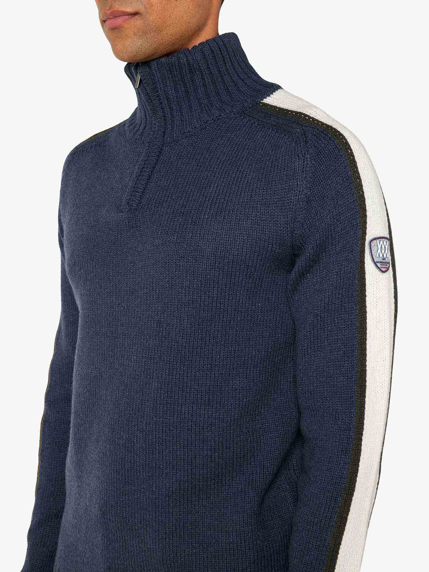 A person is wearing a navy blue Zermatt Sweater Men from We Norwegians, featuring a zippered high collar and white and black stripes running down the sleeves. This Merino Wool sweater also sports a small patch emblem on the left sleeve. The person's face is partially cropped out of the image.