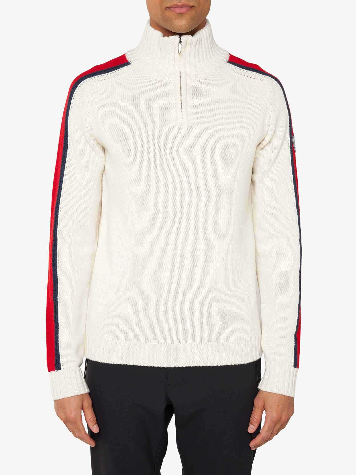 A man is dressed in a white We Norwegians Zermatt Sweater Men, which boasts a half-zip design and long sleeves adorned with striking red and black stripes extending from shoulder to wrist. Made from luxurious Merino wool, this sweater is perfectly complemented by sleek black pants for an effortlessly stylish appearance.