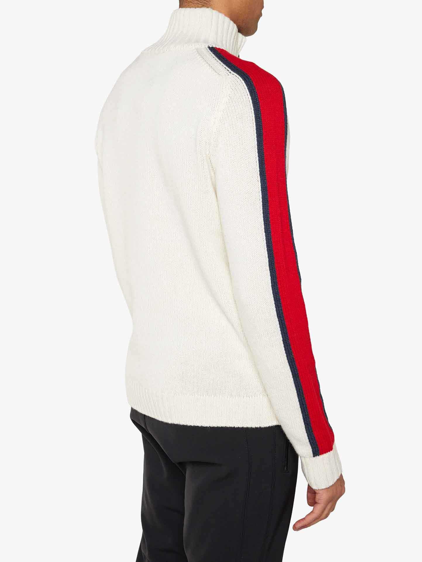 A person is wearing a We Norwegians Zermatt Sweater Men, featuring red and navy blue stripes running along the length of each sleeve. The Merino wool garment adds a touch of elegance as they stand with their back to the camera, also dressed in black pants. The background is plain white.