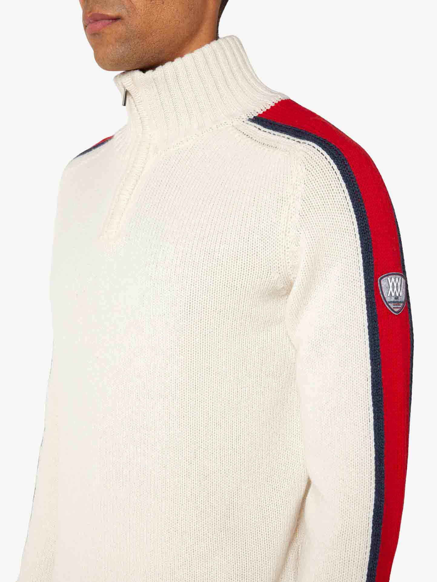 A person is wearing the We Norwegians Zermatt Sweater Men in a cream color with a high collar. The Merino wool garment features red and navy stripes running down the sleeves, along with a patch bearing an emblem on the upper arm. The background is plain white.