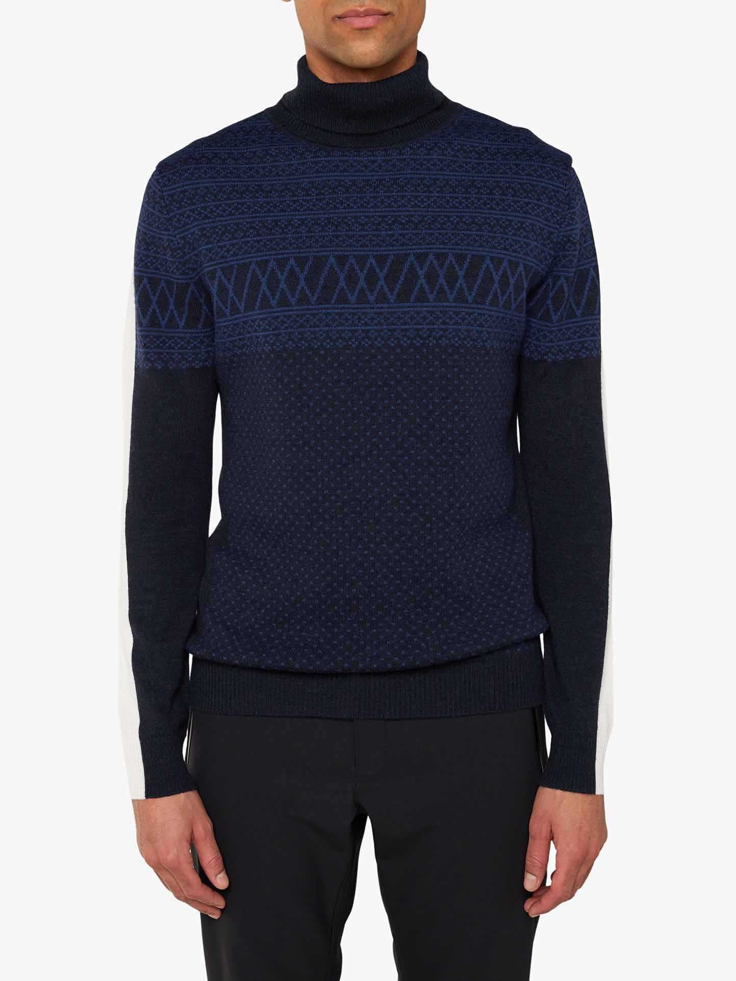 A person is wearing the "Signature Rollneck Men" sweater by We Norwegians. This dark navy blue sweater features intricate geometric Jacquard patterns in lighter blue across the chest and upper arms. It has a high, black roll neck, dark gray sleeves with white sections on the outer forearms, and is paired with black pants.