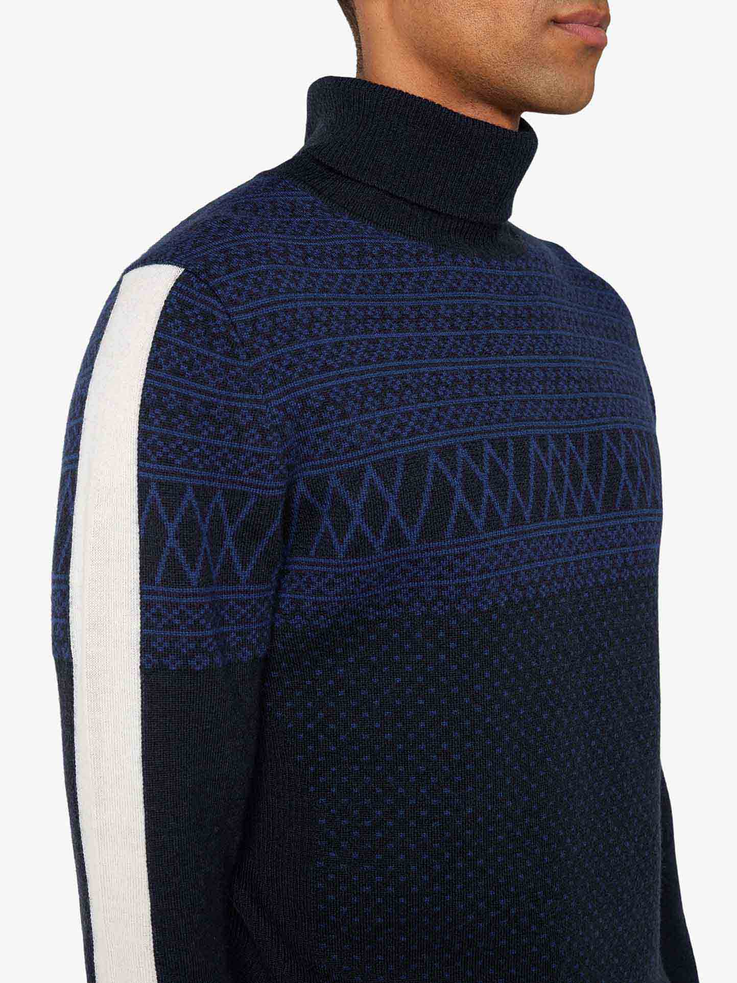 A person is shown from the shoulder up, wearing the We Norwegians Signature Rollneck Men sweater, which features intricate blue jacquard patterns and a white stripe along the sleeve. The image captures the right side profile, with the person's face partially out of frame.