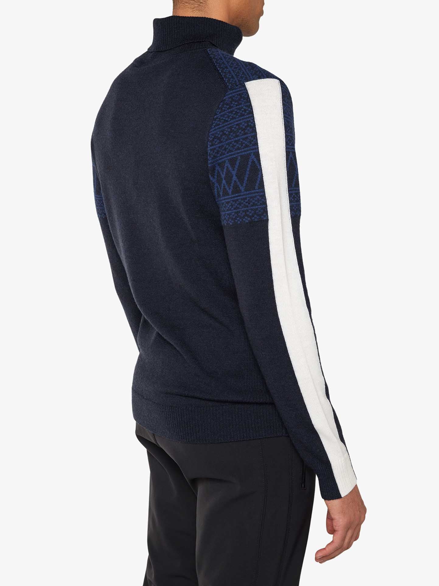 A person is seen from behind wearing the Signature Rollneck Men sweater by We Norwegians, which is navy blue with a white stripe along each sleeve. The Merino wool sweater features a knit pattern on the upper arms and shoulders. The person is also wearing black pants.