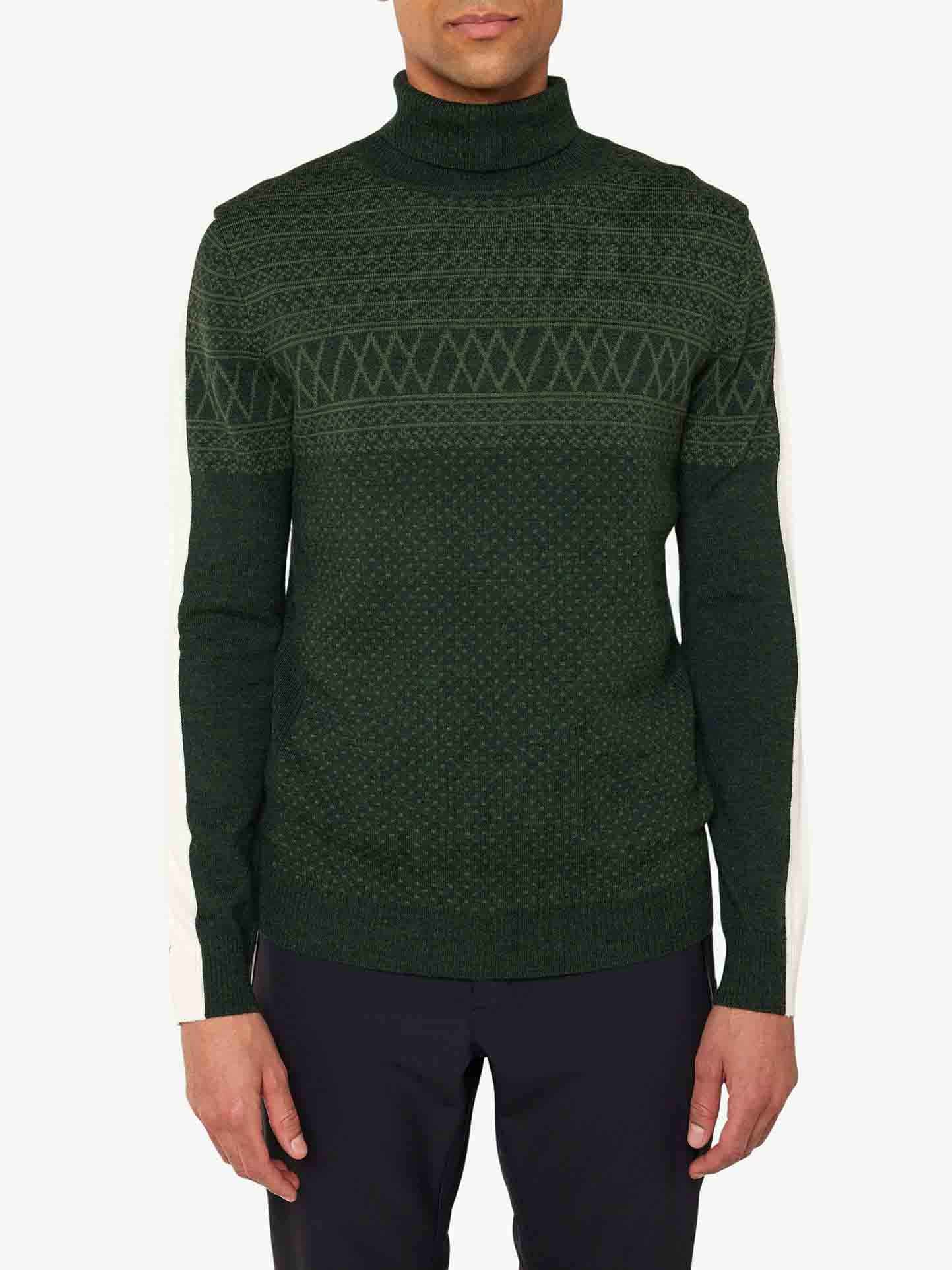 Person wearing the Signature Rollneck Men from We Norwegians, featuring a dark green hue with a jacquard pattern and white sleeves, paired with dark trousers, standing against a white background.