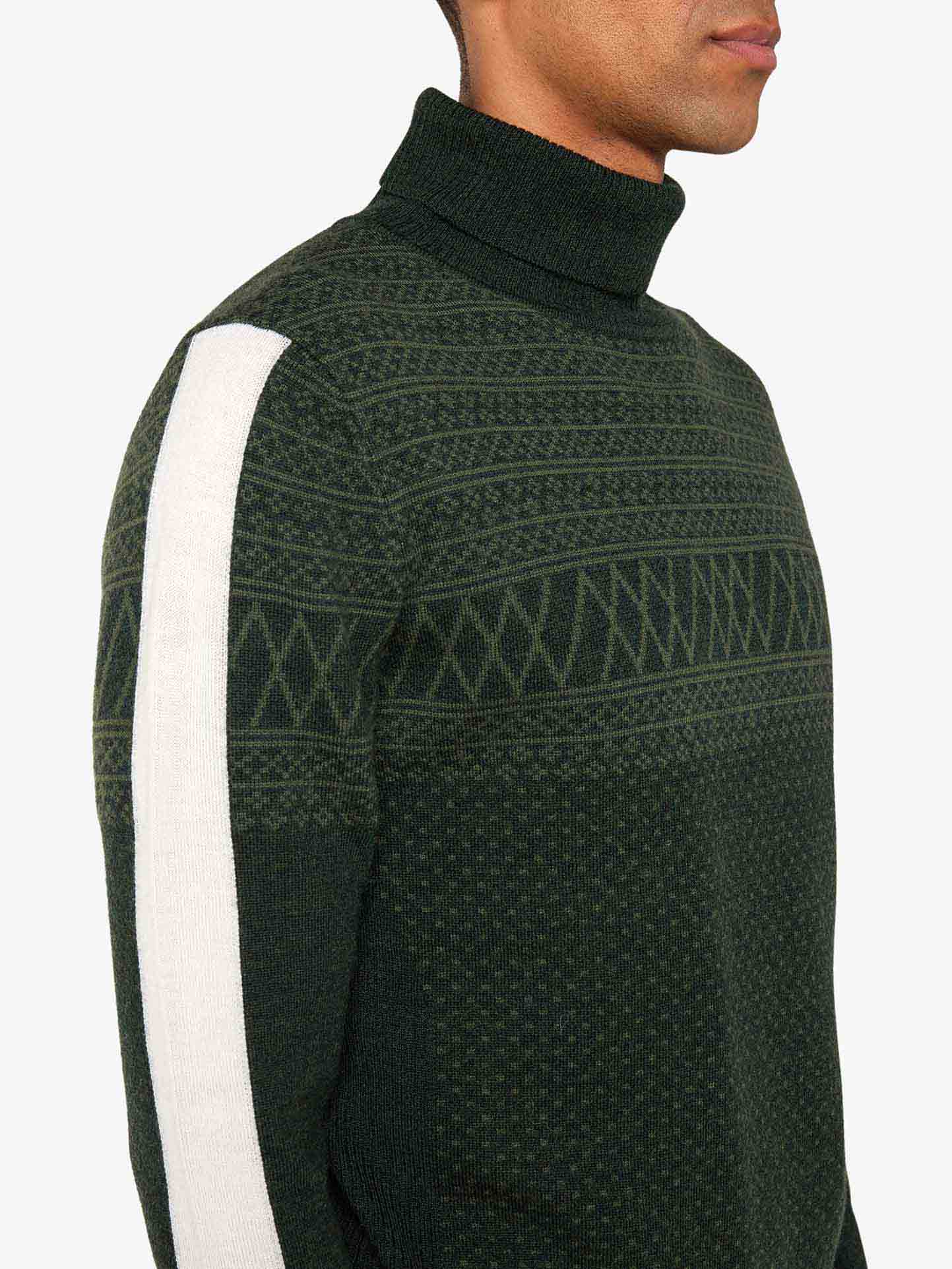 A person wearing the "Signature Rollneck Men" sweater by We Norwegians. The dark green merino wool sweater features a high roll neck collar, intricate jacquard patterns, and a white stripe running along the length of the sleeve. The person's head is cropped out of the image.