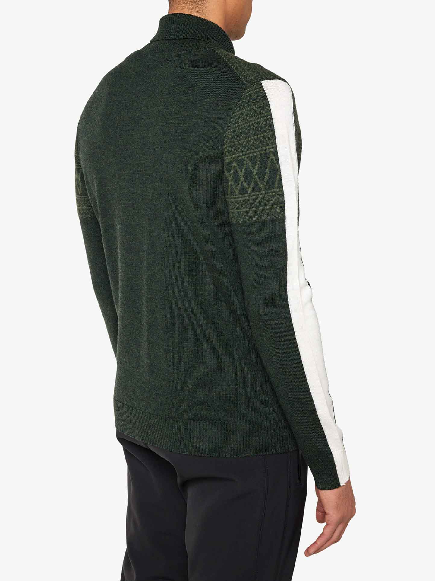 A person is wearing the Signature Rollneck Men sweater by We Norwegians, a dark green Merino wool roll neck featuring a white stripe and Jacquard pattern on the upper part of the right sleeve. They are also dressed in dark pants against a plain white background.