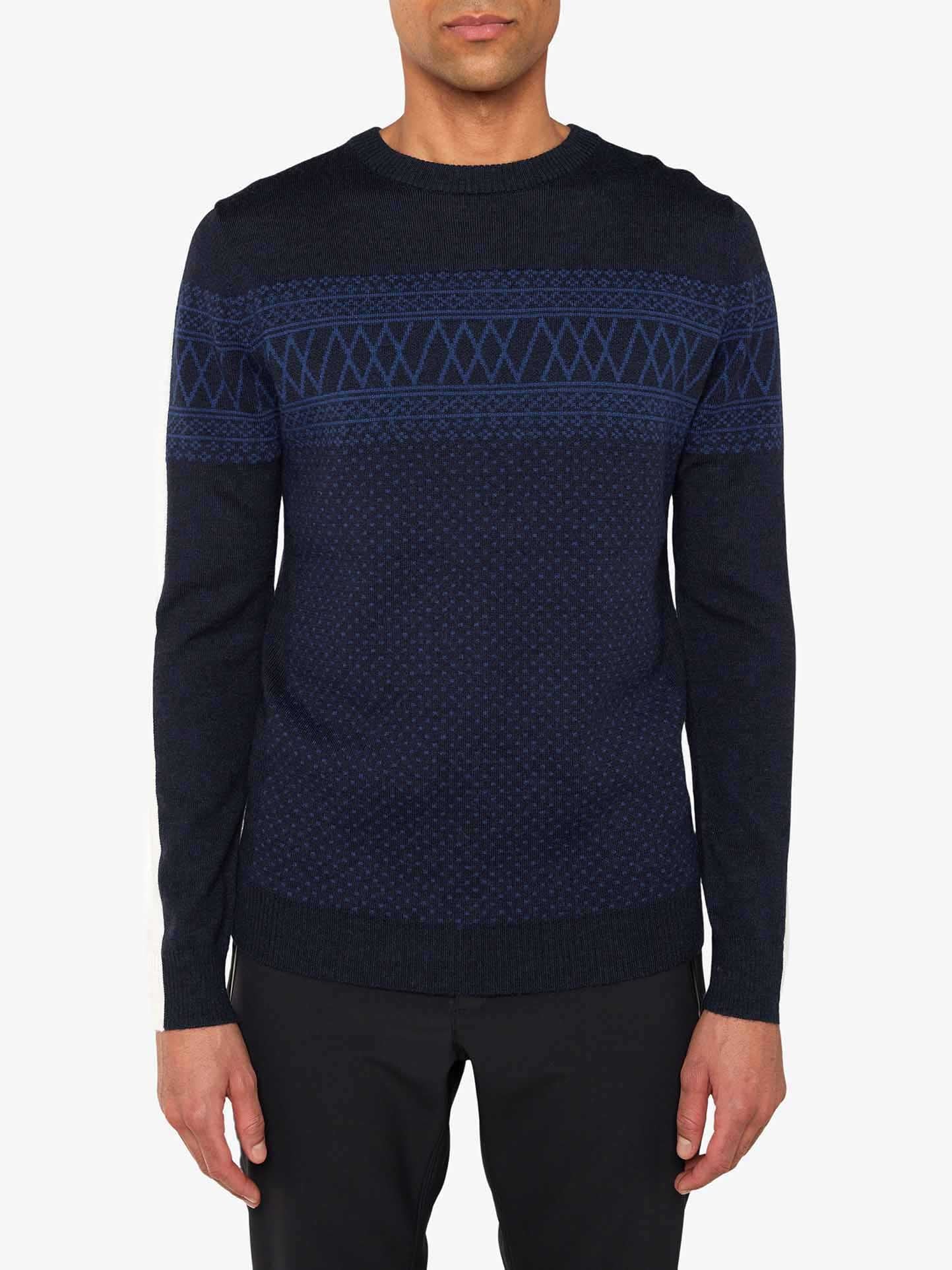 A person is wearing the We Norwegians Signature Crewneck Men sweater in navy blue, featuring a slim fit and geometric Setesdal pattern with diamond shapes and horizontal lines across the chest, intricately knitted with washable merino yarn. The outfit is completed with black pants, against a plain white background.