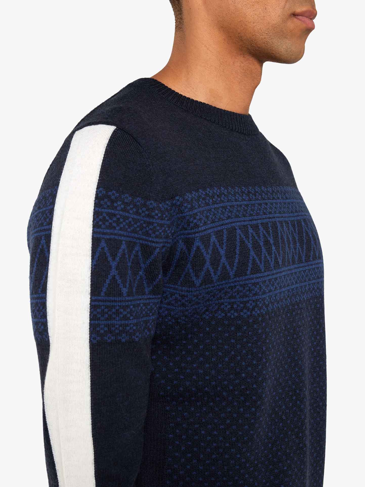 A person is shown from the side wearing the We Norwegians Signature Crewneck Men, a slim-fit dark blue sweater with intricate light blue Setesdal patterns on the chest and sleeves. The sweater also features a prominent white stripe running down the length of the outer sleeve and is made from washable merino yarn. The background is plain white.