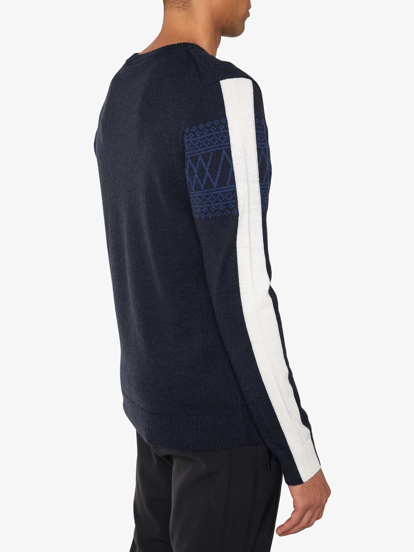 A person wearing the We Norwegians Signature Crewneck Men in dark blue, which features a white stripe running down the length of one sleeve and a subtle blue geometric Setesdal pattern on the upper part of the sleeve. Made from washable merino yarn, they are facing away from the camera, showcasing the back of this slim fit sweater.