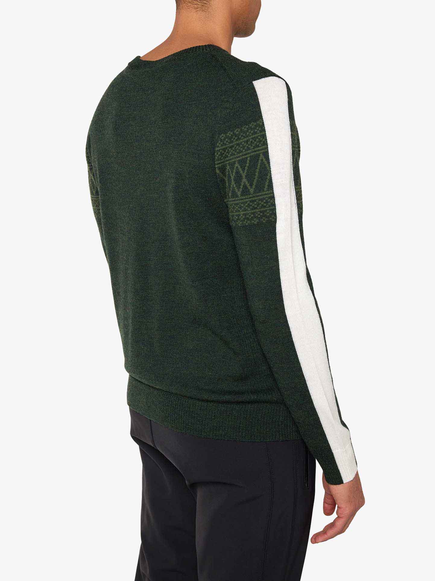 A person wearing the Signature Crewneck Men by We Norwegians—in a dark green, washable merino yarn with white patterned detailing on the sleeves—and black pants is standing with their back to the camera. The slim-fit sweater showcases a white vertical stripe on the left arm and geometric patterns near the shoulder.