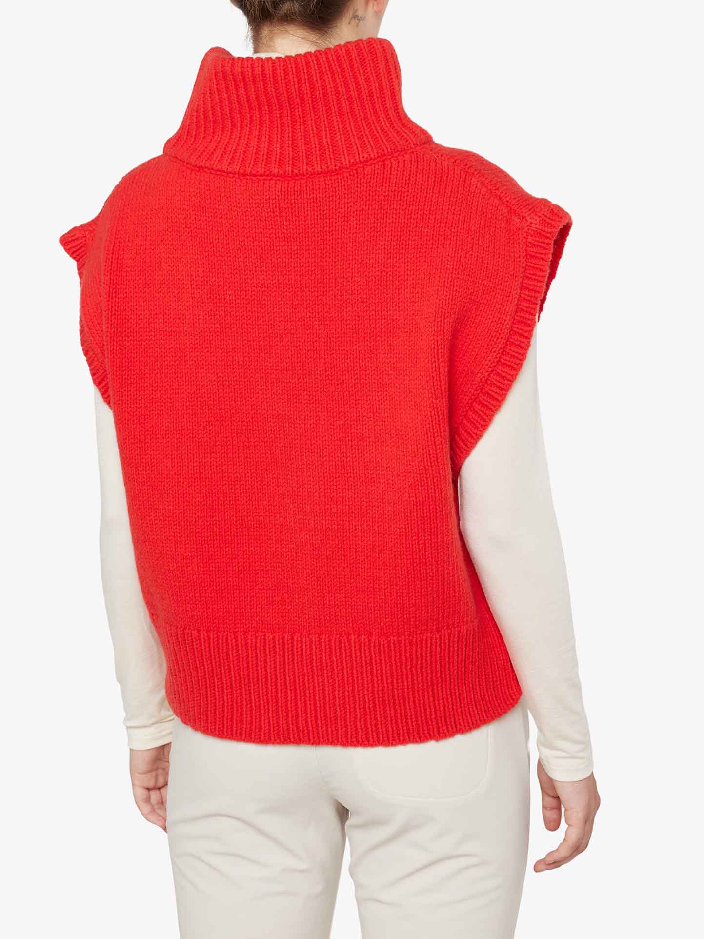 A person is seen from the back, wearing the cozy and warm Setesdal Vest Women by We Norwegians in a red turtleneck sleeveless style over a long-sleeve white top. The vest, crafted with merino/cashmere yarn, features a thick, ribbed neckline and hem. The person's hair is tied up, and they are dressed in light-colored pants.