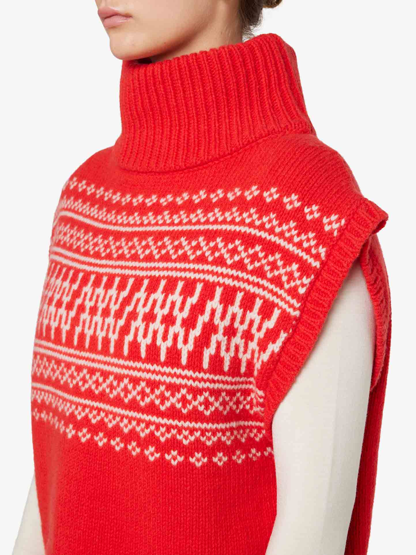 A person wearing the Setesdal Vest Women by We Norwegians, a vibrant red, sleeveless turtleneck sweater crafted from cozy warm merino/cashmere yarn and featuring a white geometric pattern across the chest. The vest is layered over a long-sleeve white top. The photo is taken from the side, focusing on the upper torso and neck.