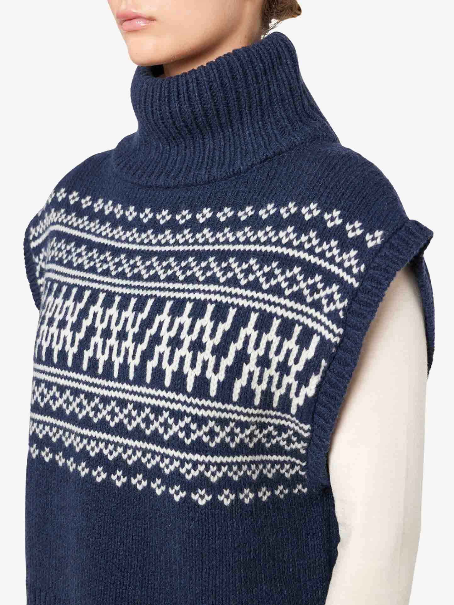 The individual is wearing a sleeveless, navy blue Setesdal Vest Women by We Norwegians, featuring a white geometric Fair Isle pattern across the chest and shoulders. This cozy and warm vest is crafted from luxurious merino/cashmere yarn. The person stands against a plain background, with the focus on the upper body and sweater details.