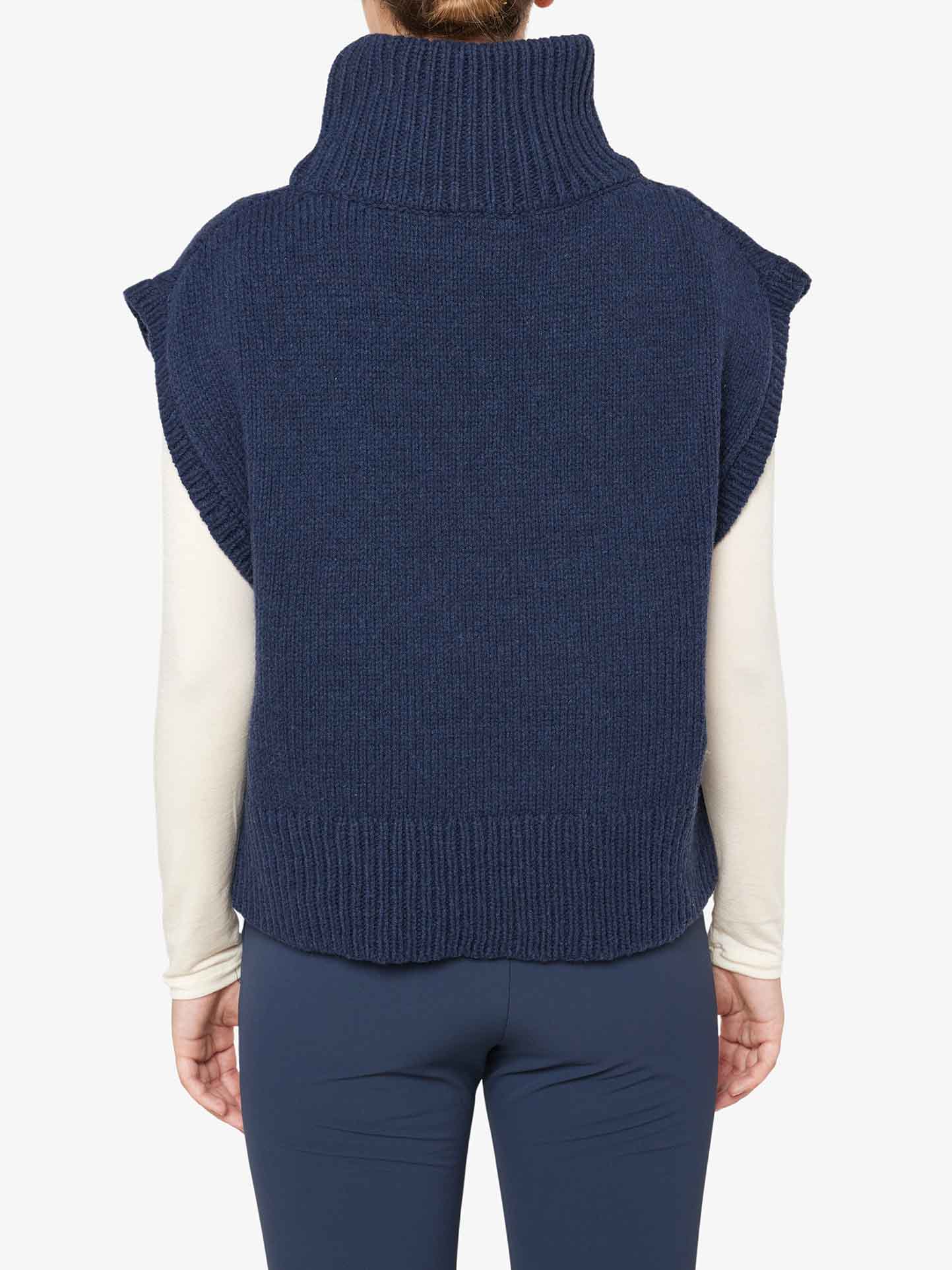 A person is shown from the back, wearing the We Norwegians Setesdal Vest Women – a dark blue, sleeveless turtleneck sweater crafted from merino/cashmere yarn. It is layered over a white long-sleeve shirt and paired with dark pants. The cozy, warm vest features a ribbed texture at the collar, armholes, and hem.