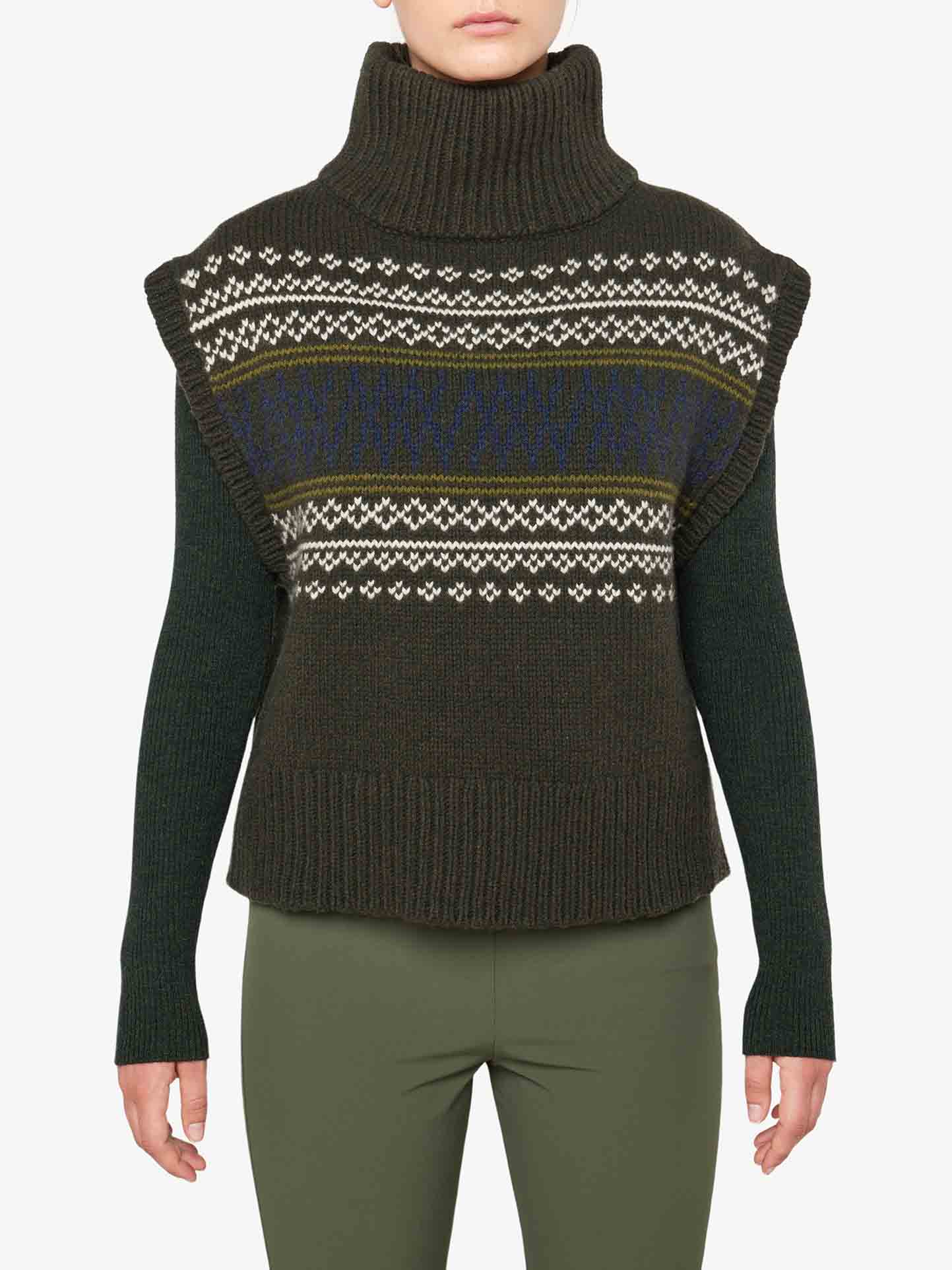 A person is wearing the Setesdal Vest Women from We Norwegians, a high-neck, sleeveless dark green sweater featuring Norwegian knits across the chest. It is layered over a long-sleeve dark green top and paired with olive green pants. Their face is not visible in the image.