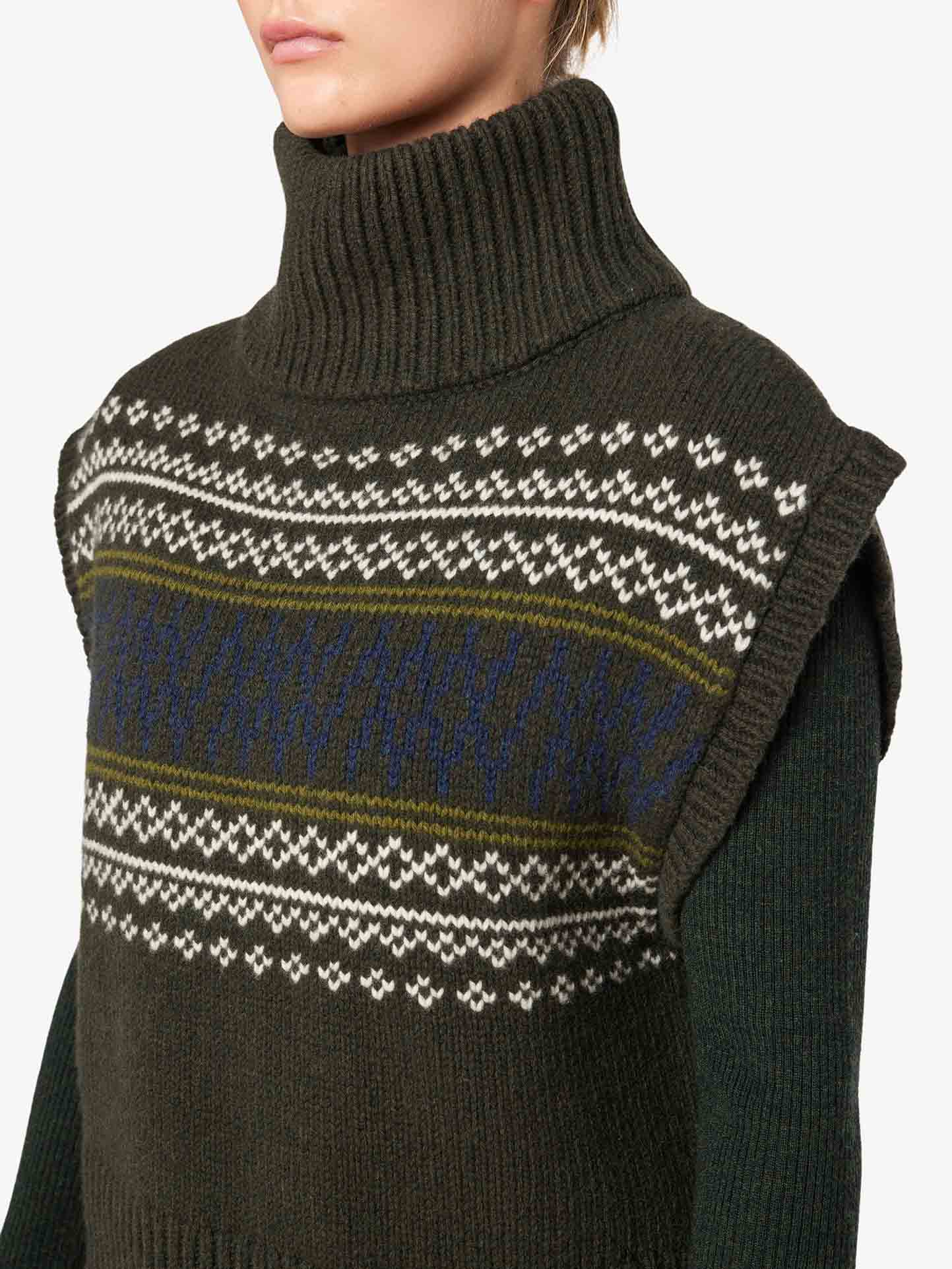 A person is wearing the Setesdal Vest Women from We Norwegians, a dark green turtleneck sweater with a knitted pattern reminiscent of Norwegian knits. The pattern features horizontal stripes of white and blue designs across the chest and upper sleeves. The image is cropped to focus on the upper torso and neck, without showing the person's face.