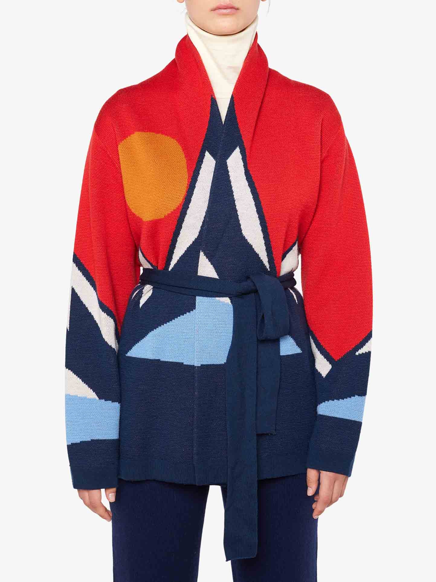 A person wearing the vibrant Snowmountain Cardigan Women by We Norwegians, featuring abstract geometric patterns in red, blue, white, and orange tones. Made from luxurious Merino wool, the cardigan has long sleeves and a belt tied around the waist. The person also wears a white turtleneck sweater underneath, creating an après-ski look.