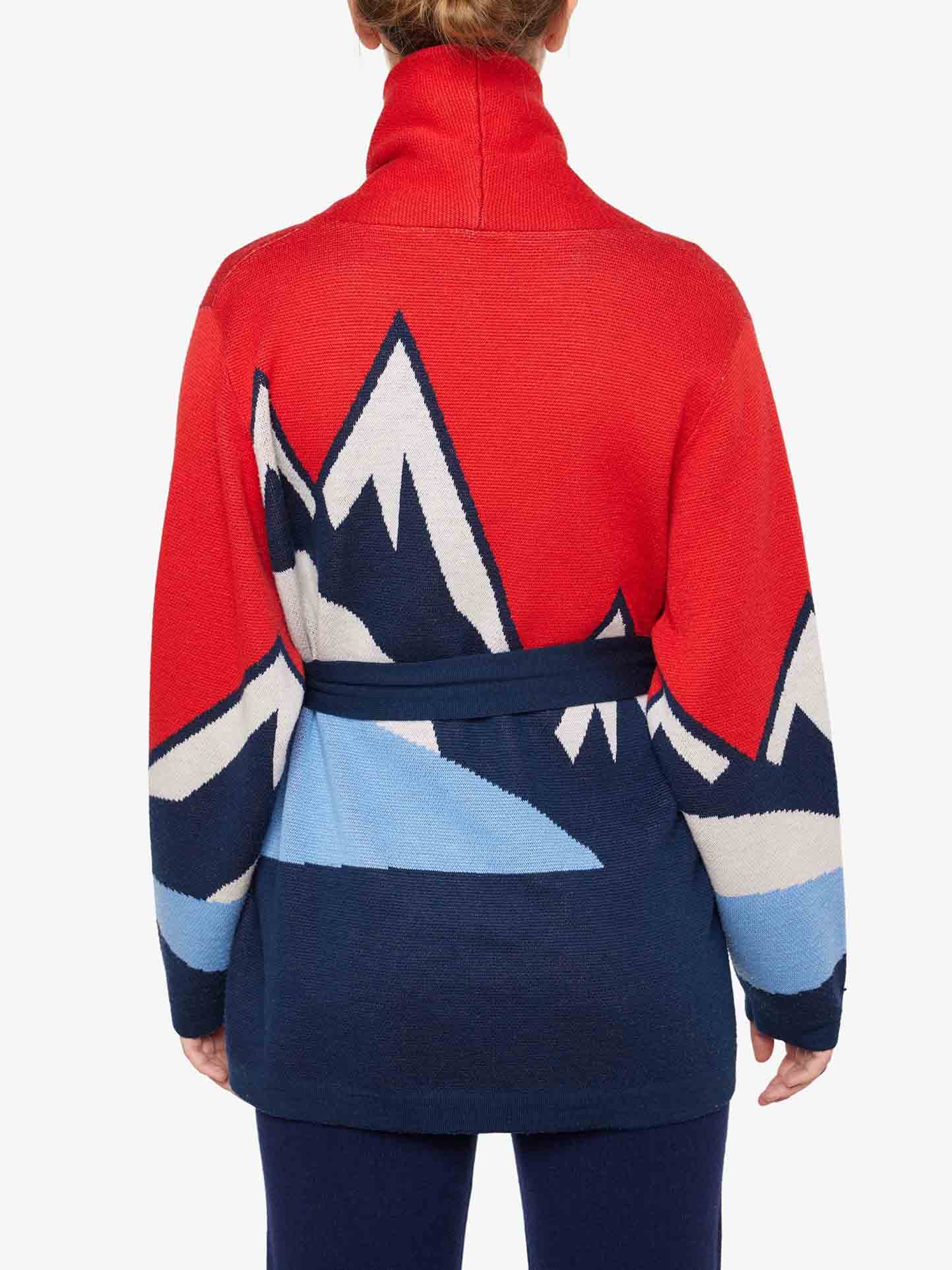 A person wearing a Snowmountain Cardigan Women by We Norwegians, characterized by its red, blue, and white Merino wool with a mountain design on the back. The belted waist and high collar enhance the après ski look. The person is viewed from behind, showcasing the colorful and distinctive pattern of the sweater.