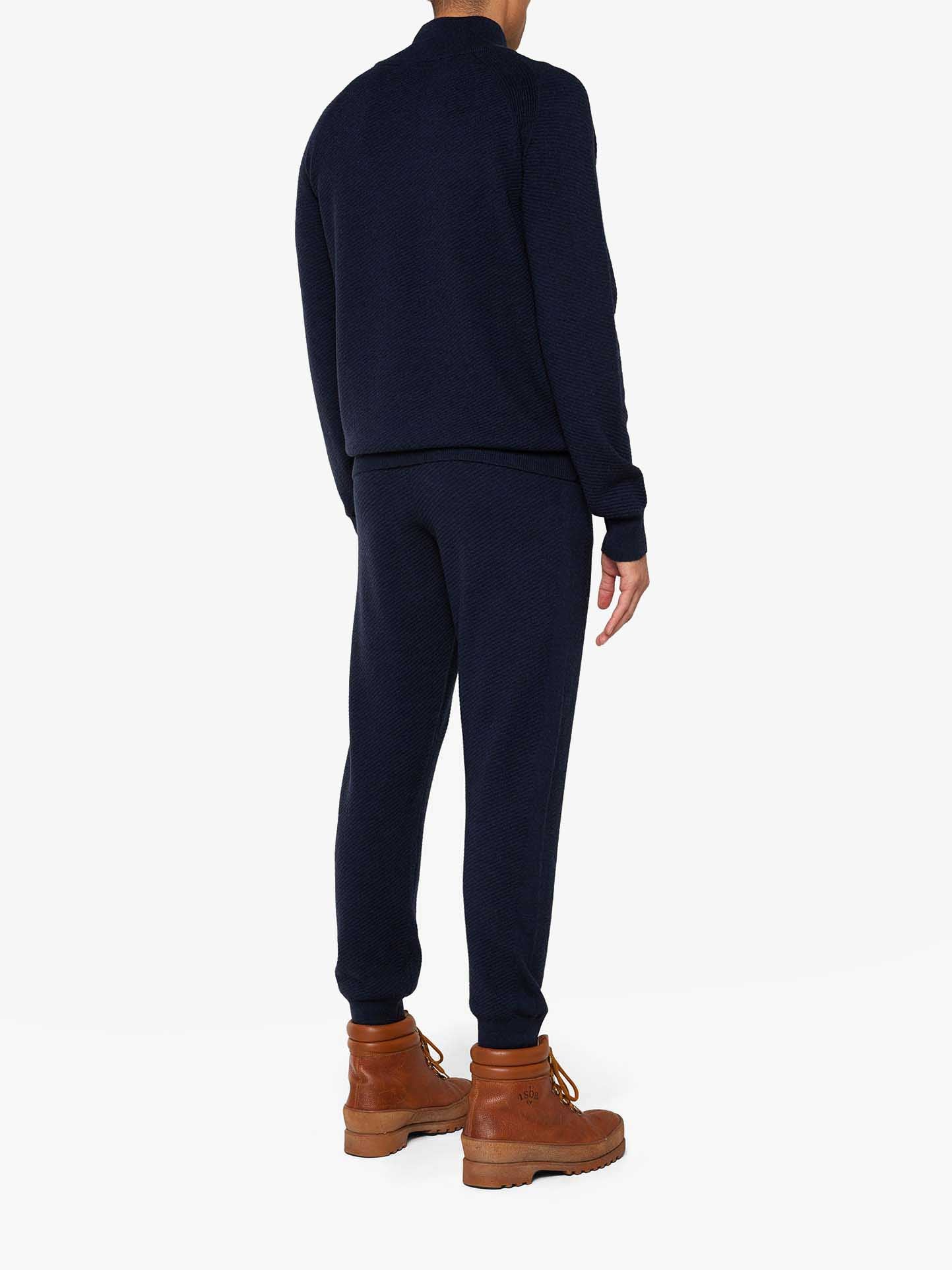 A person is standing with their back to the camera, clad in a stylish dark blue tracksuit resembling the chic Geilo 1/4-Zip Men lounge set from We Norwegians, complete with a long-sleeved top and joggers. They are also wearing brown lace-up boots, all set against a white background.